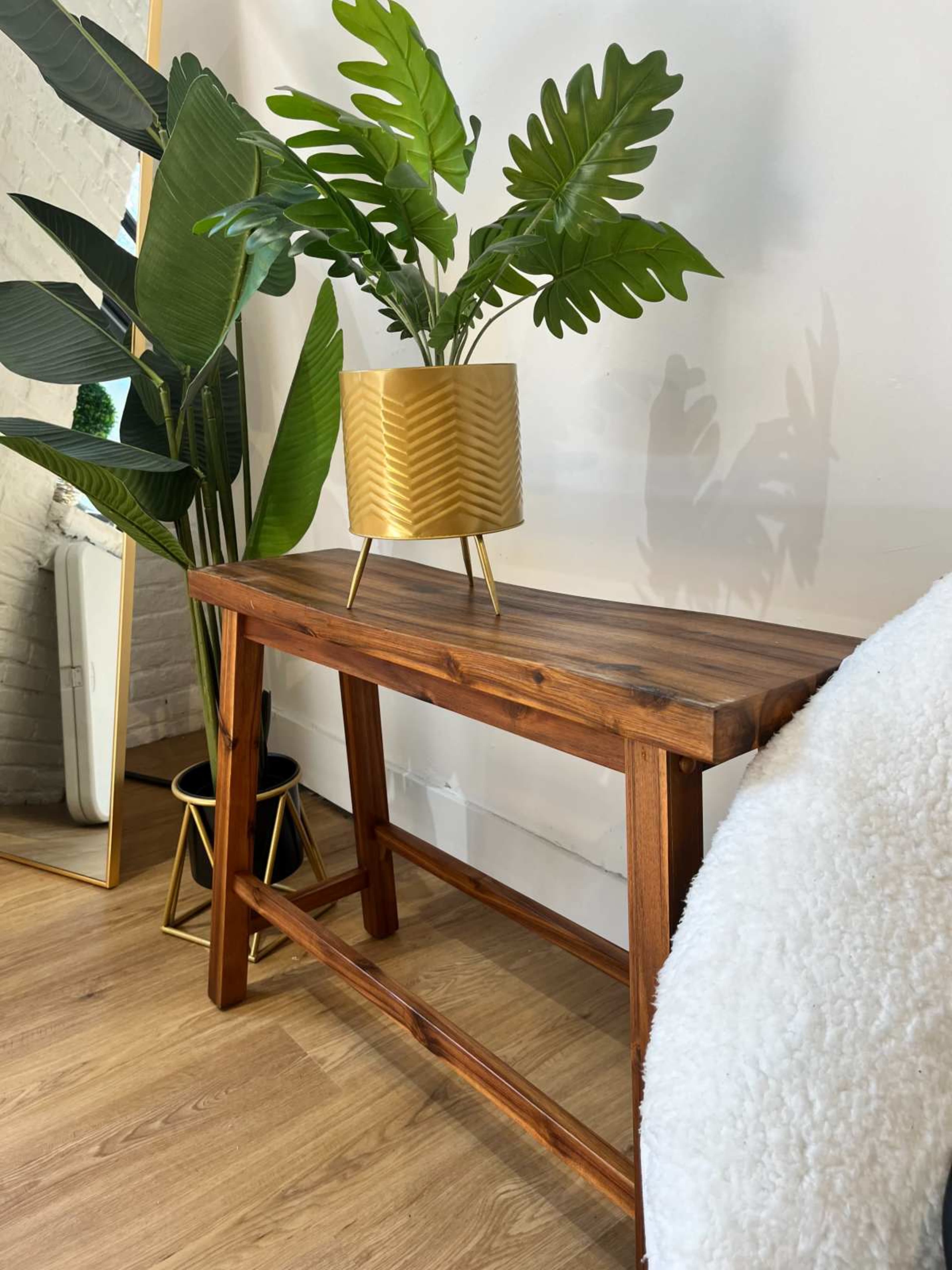 A wooden console table holds a potted plant with large green leaves, positioned beside a mirror and a tall leafy plant.