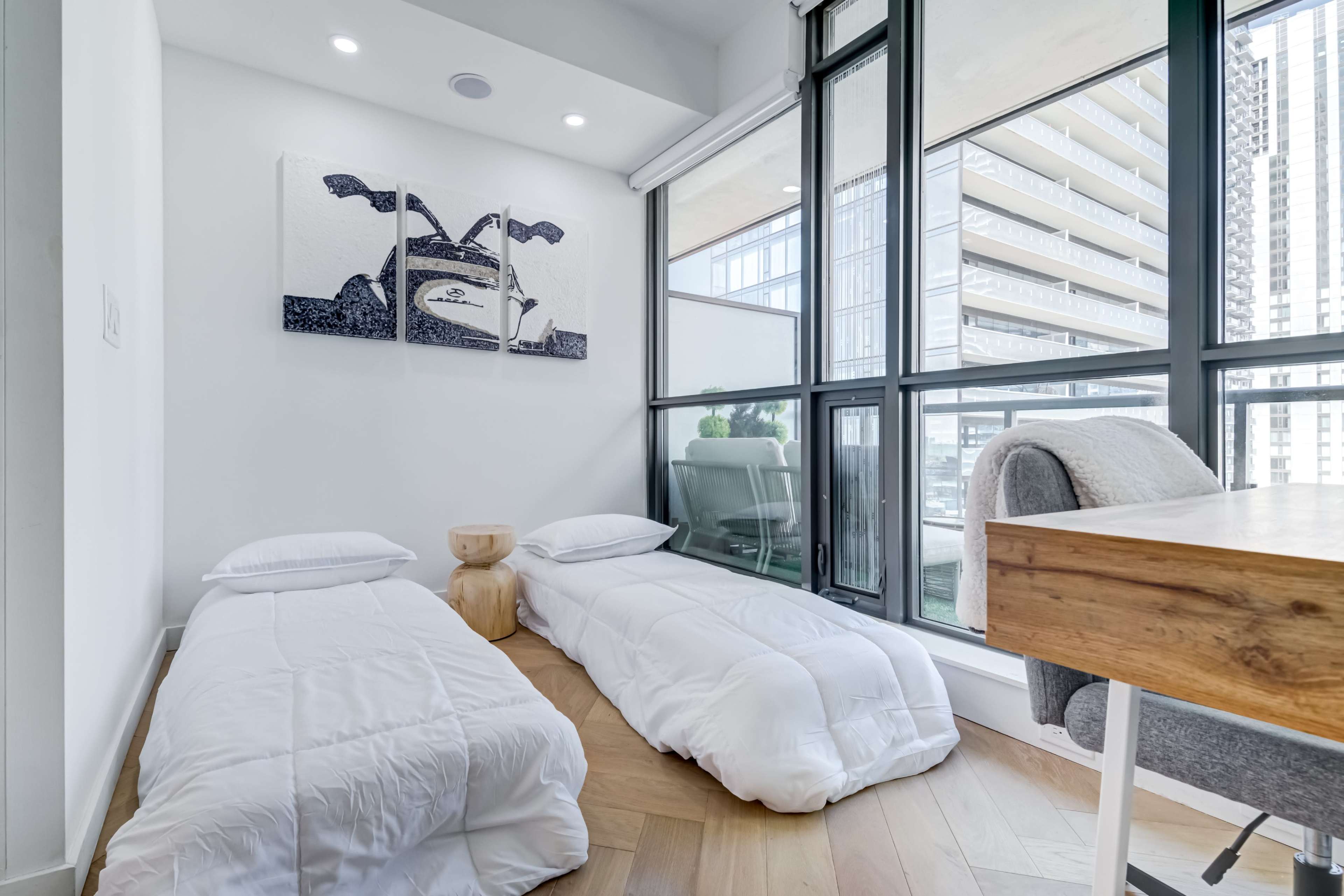 A brightly lit room features two white bedding sets on the floor beside a wooden desk and large glass windows that overlook a building facade.