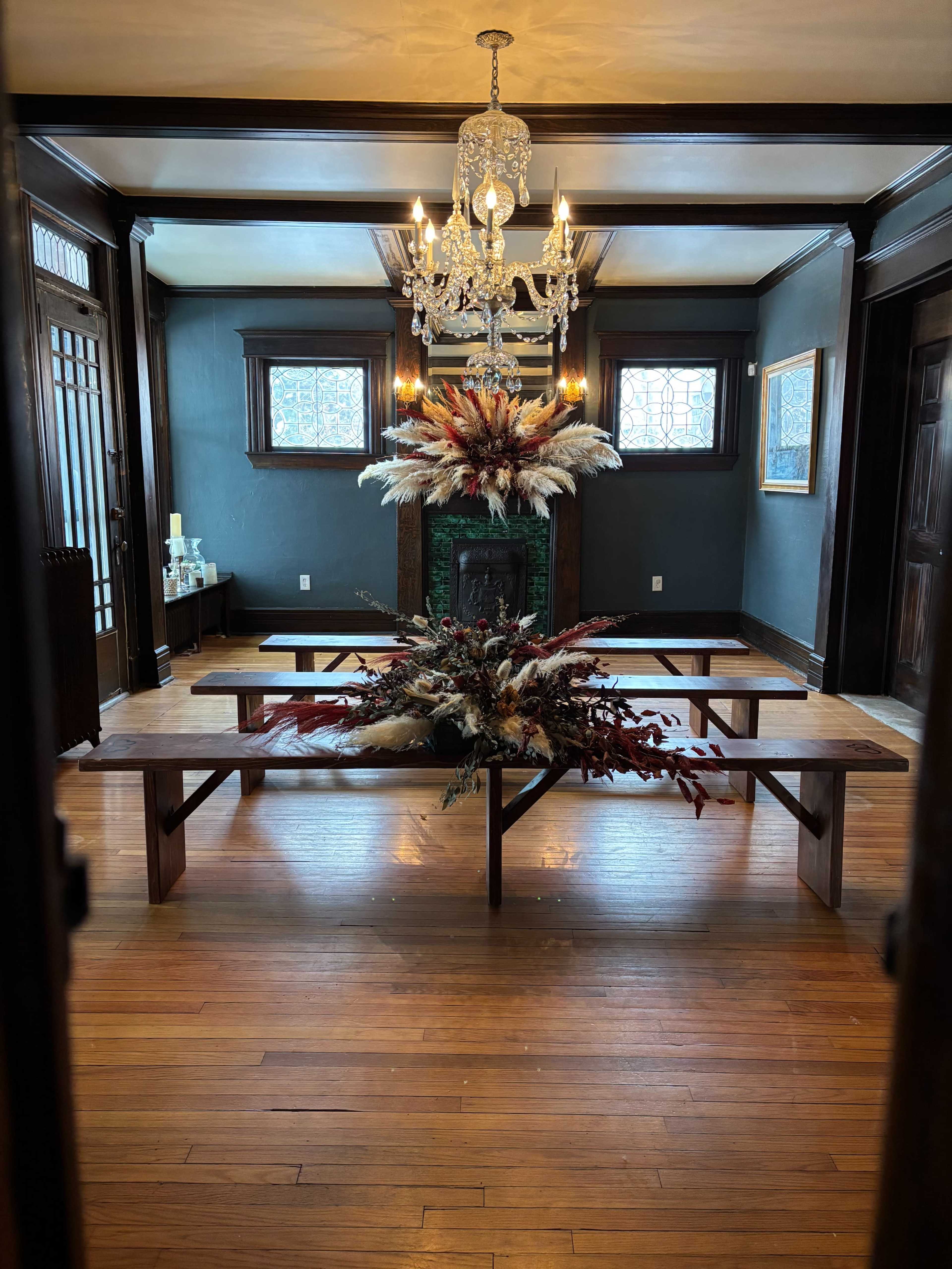 The image shows a room with dark walls, a wooden floor, a large floral arrangement on a table, and a chandelier hanging from the ceiling.