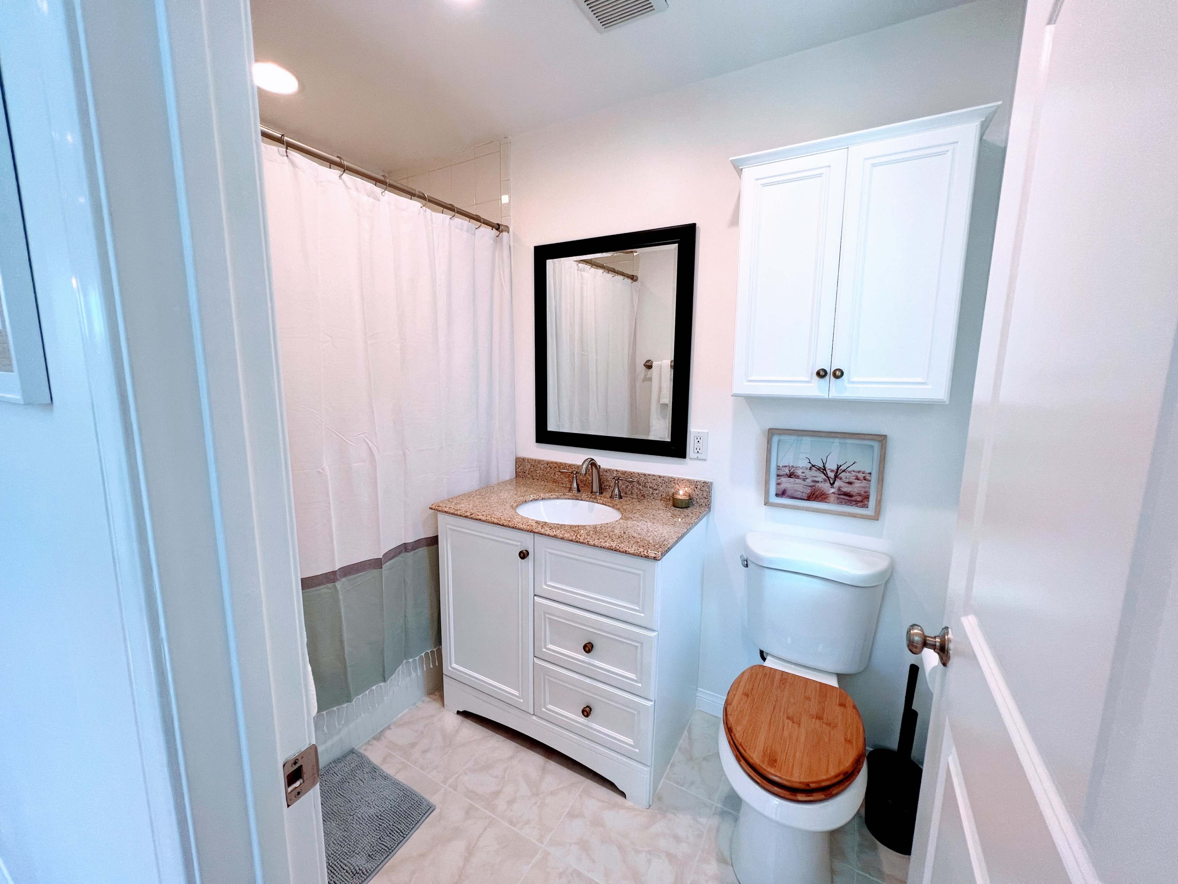 A modern bathroom features a shower with a curtain, a white vanity with a granite countertop, a light-colored tile floor, and a wooden toilet seat.