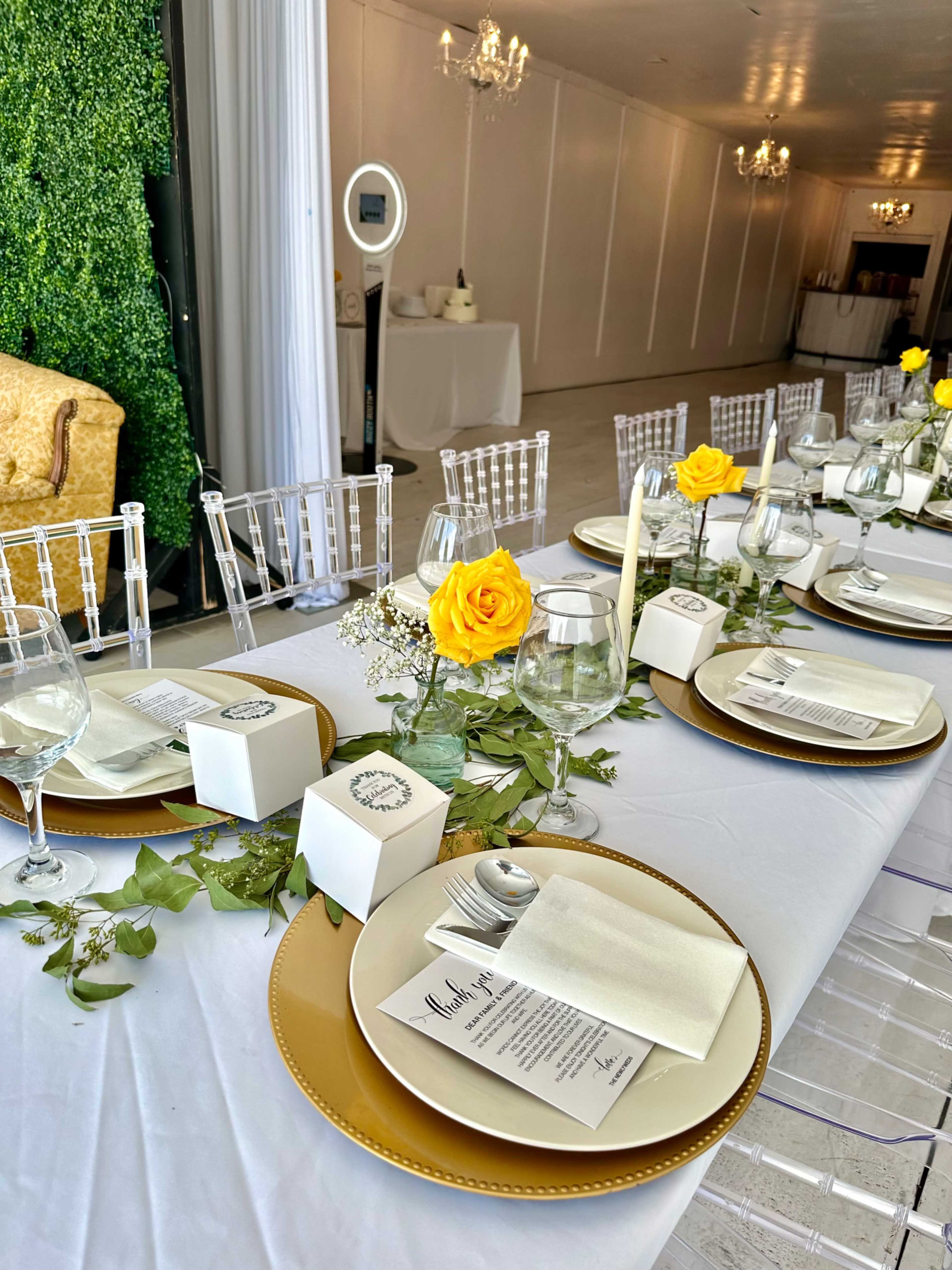 A elegantly set dining table with white tablecloths, gold chargers, glassware, and yellow roses as centerpieces.