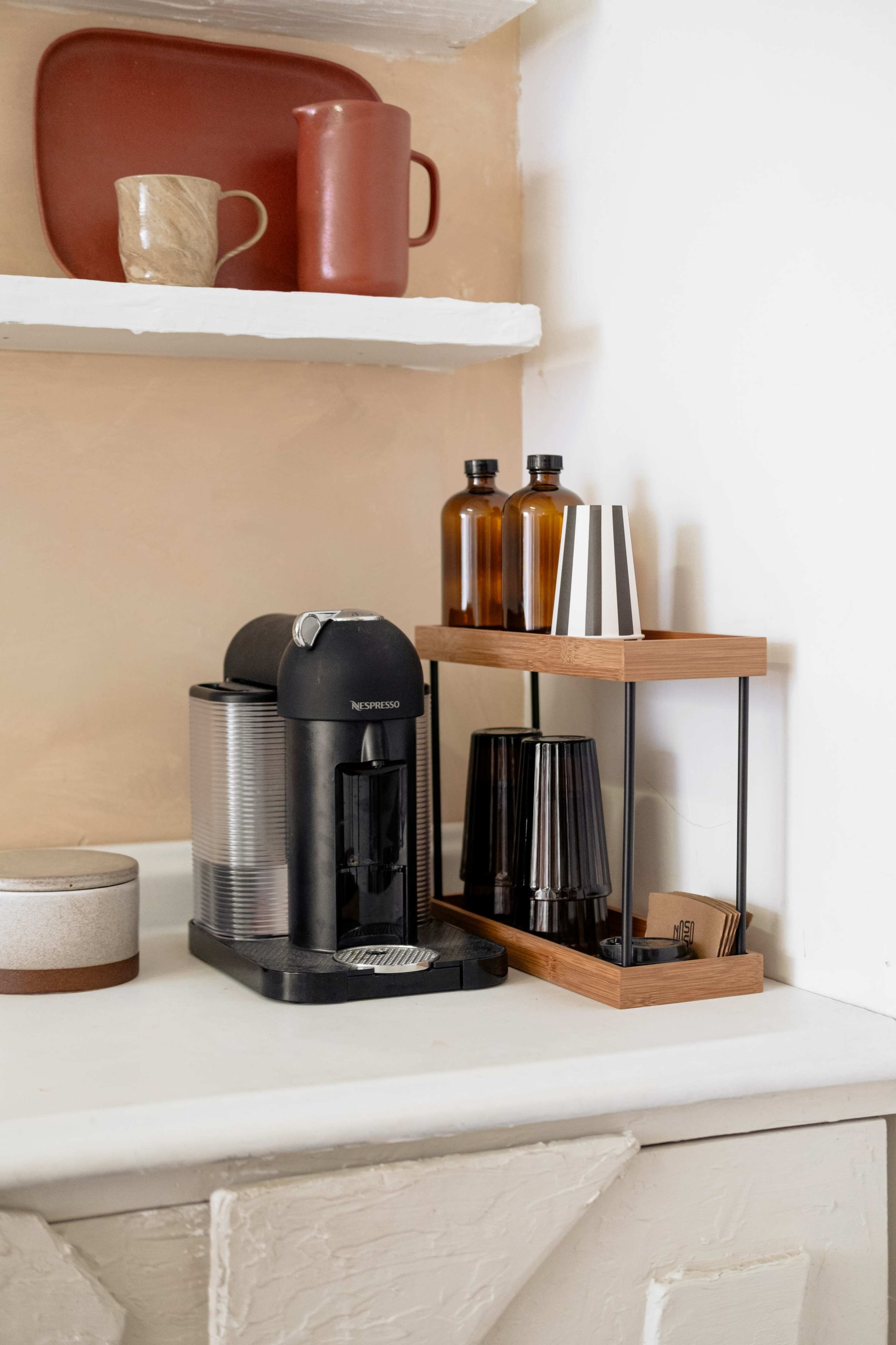 A black coffee machine sits on a white countertop next to a wooden organizer holding glasses and bottles.