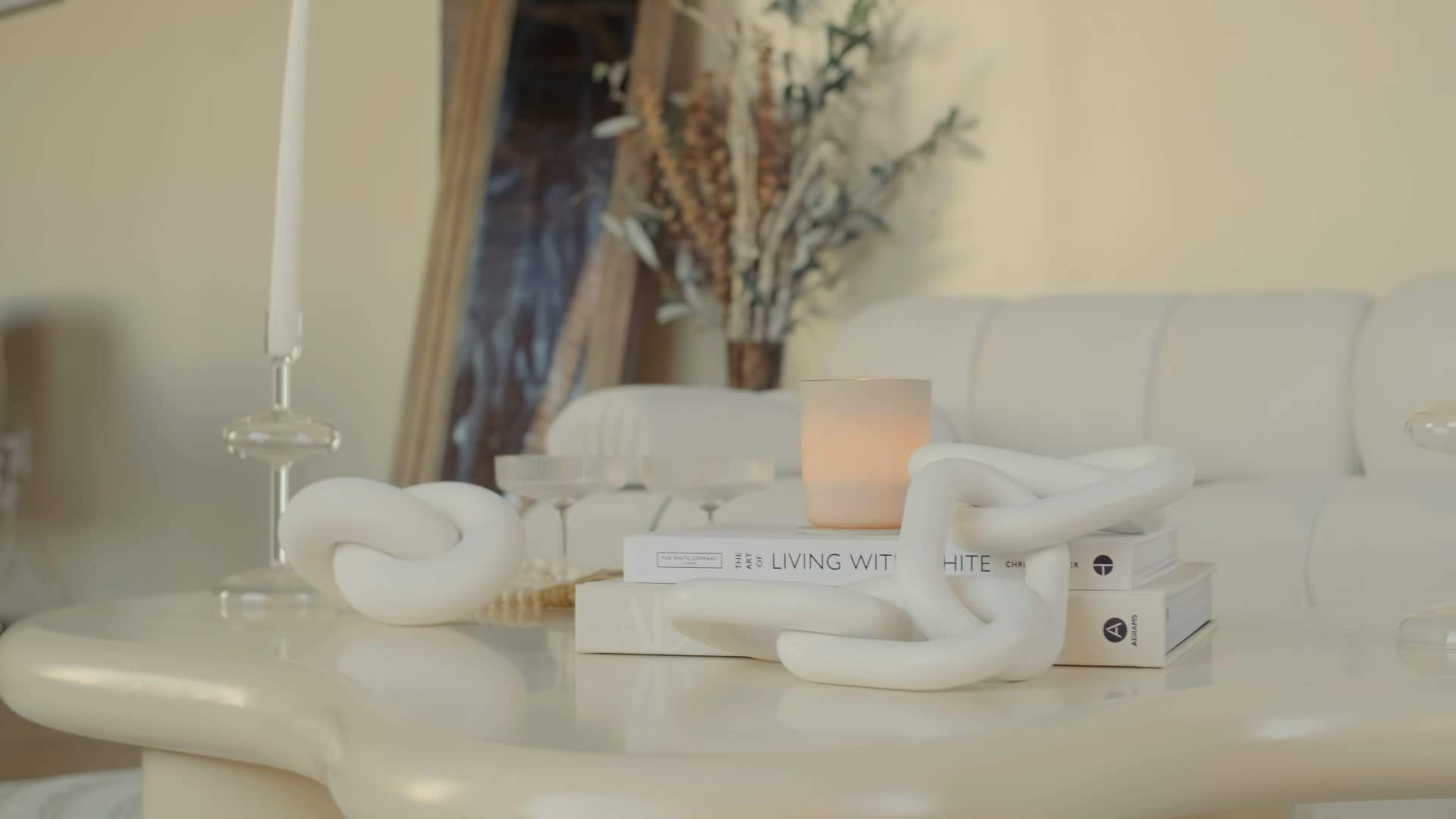 A white modern coffee table displays sculptural decor, stacked books, and a lit candle, set against a light-colored room.