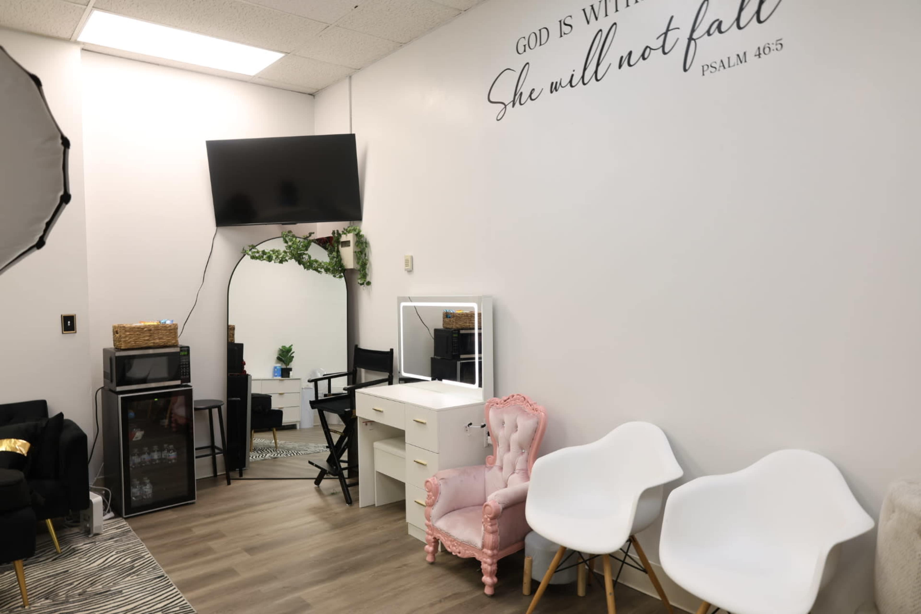 The image shows a brightly lit room featuring a pink chair, a vanity with a mirror, a TV on the wall, and modern seating arrangements.