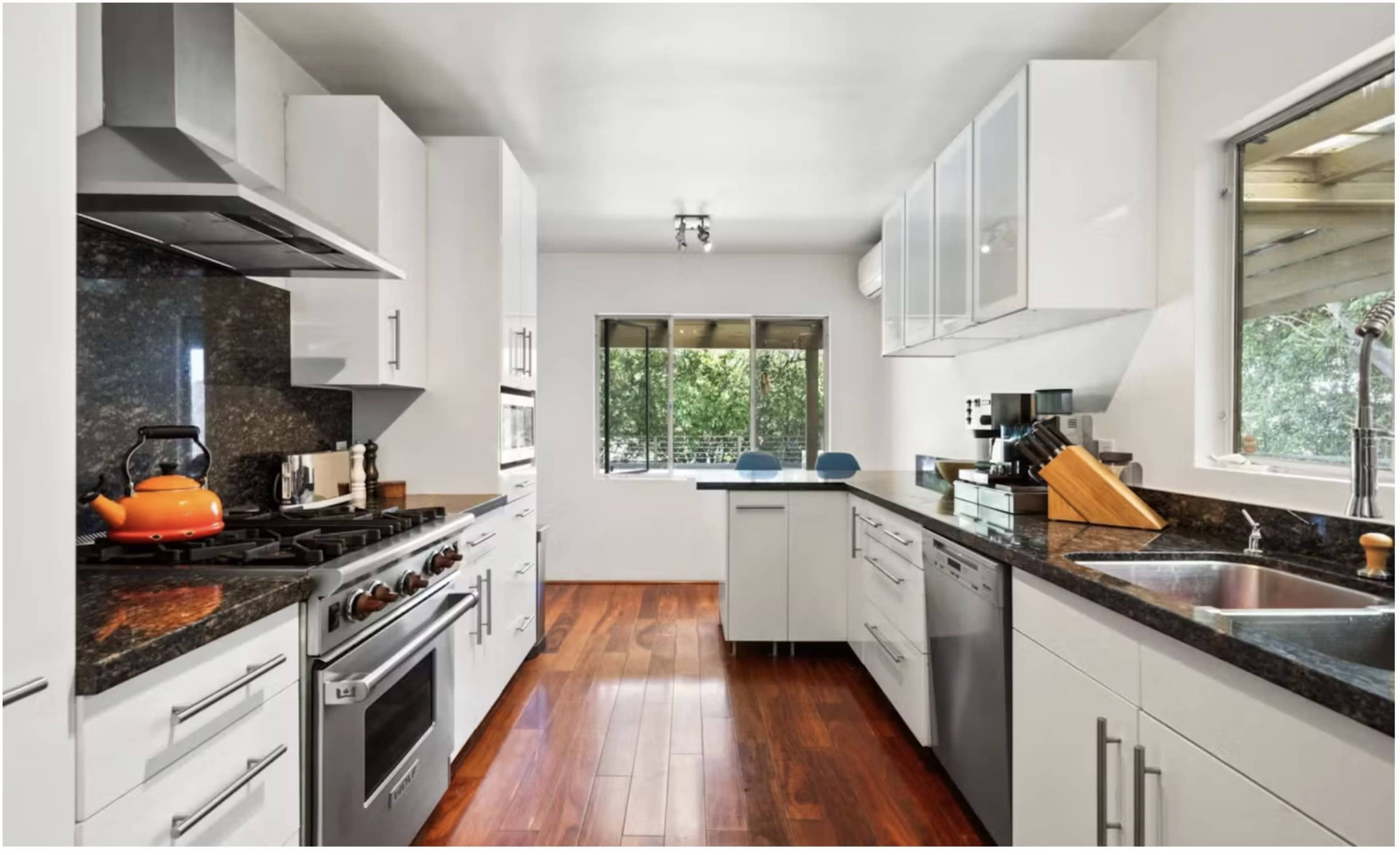 A modern kitchen features stainless steel appliances, white cabinetry, and a dark countertop, with a window showing a view of greenery outside.