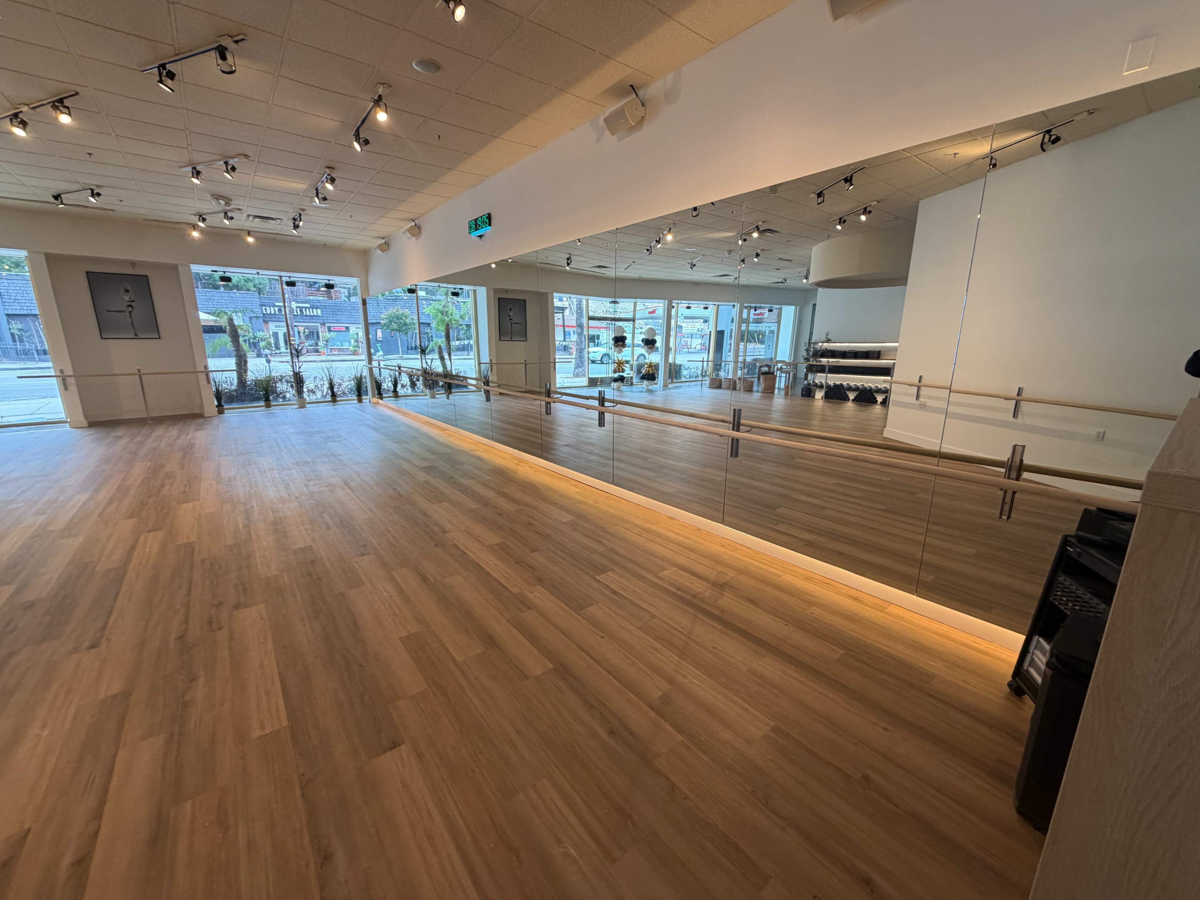 The image shows a spacious dance studio with wooden flooring, large mirrors along one wall, and natural light streaming in through windows.
