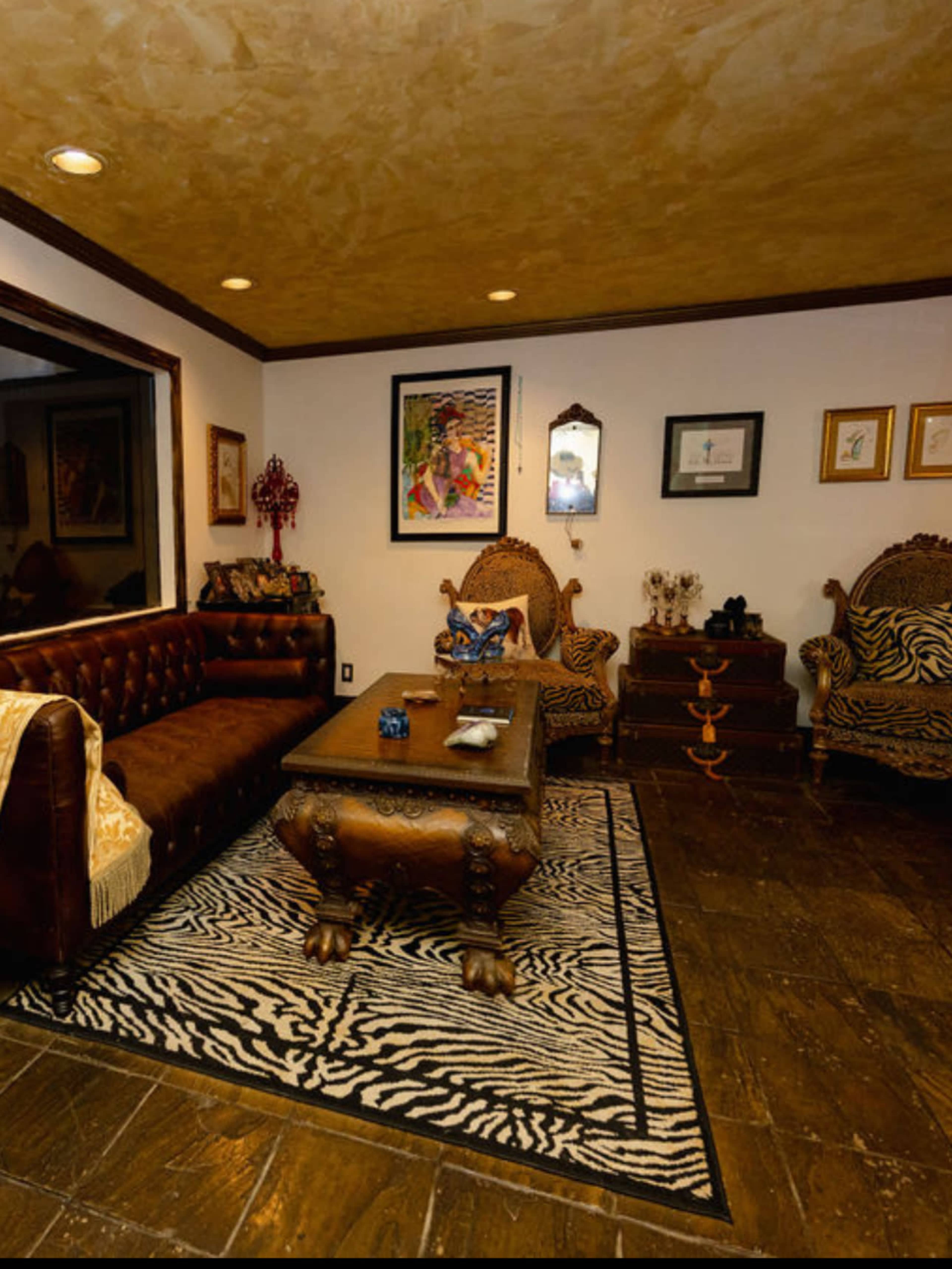 The image shows a room with a wooden floor, a brown leather sofa, ornate chairs, a zebra-striped rug, and decorated walls featuring various framed artworks.