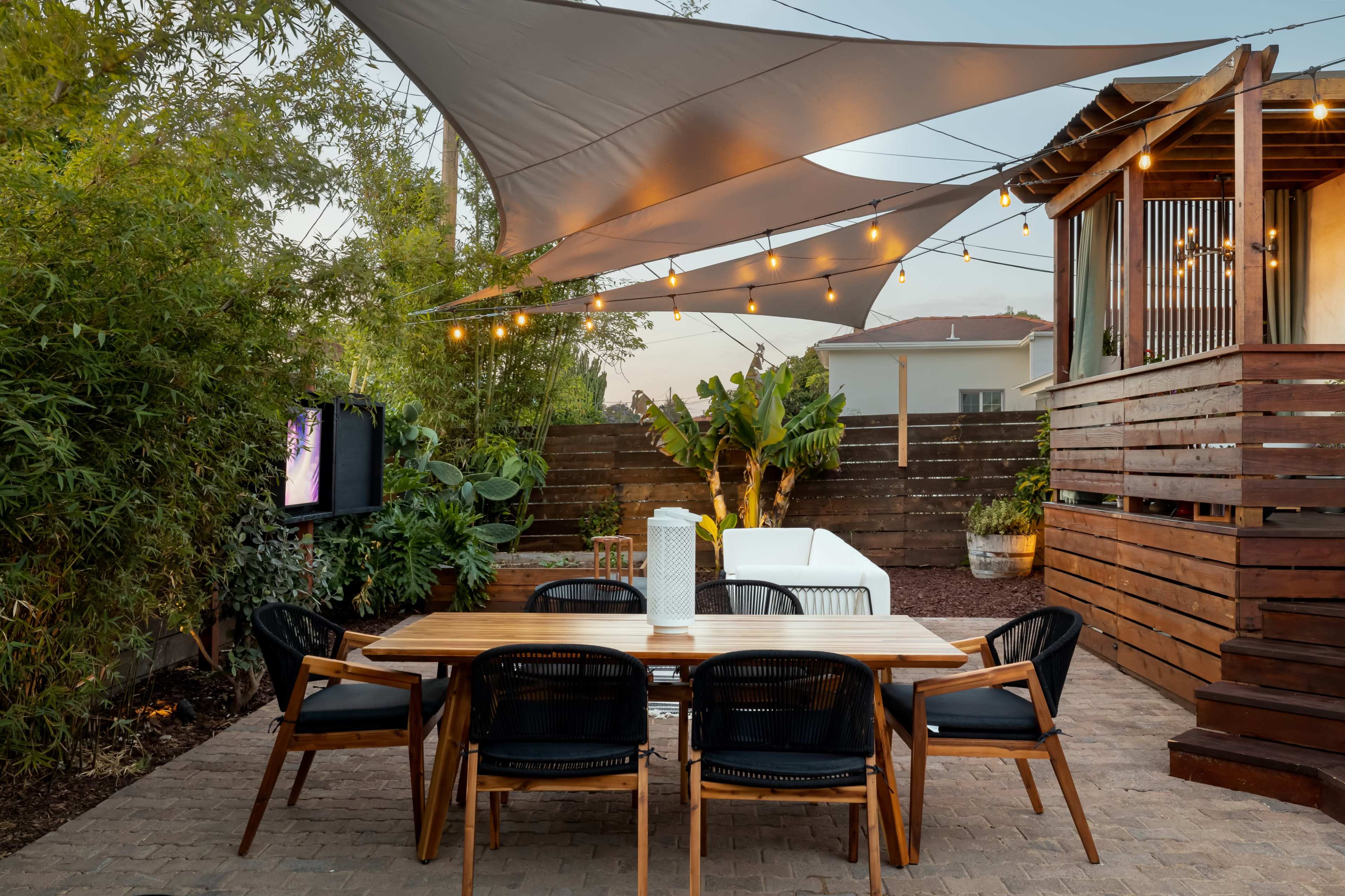 A dining table with black chairs is set up under a large shade sail, surrounded by greenery and string lights, next to a wooden deck and a television.