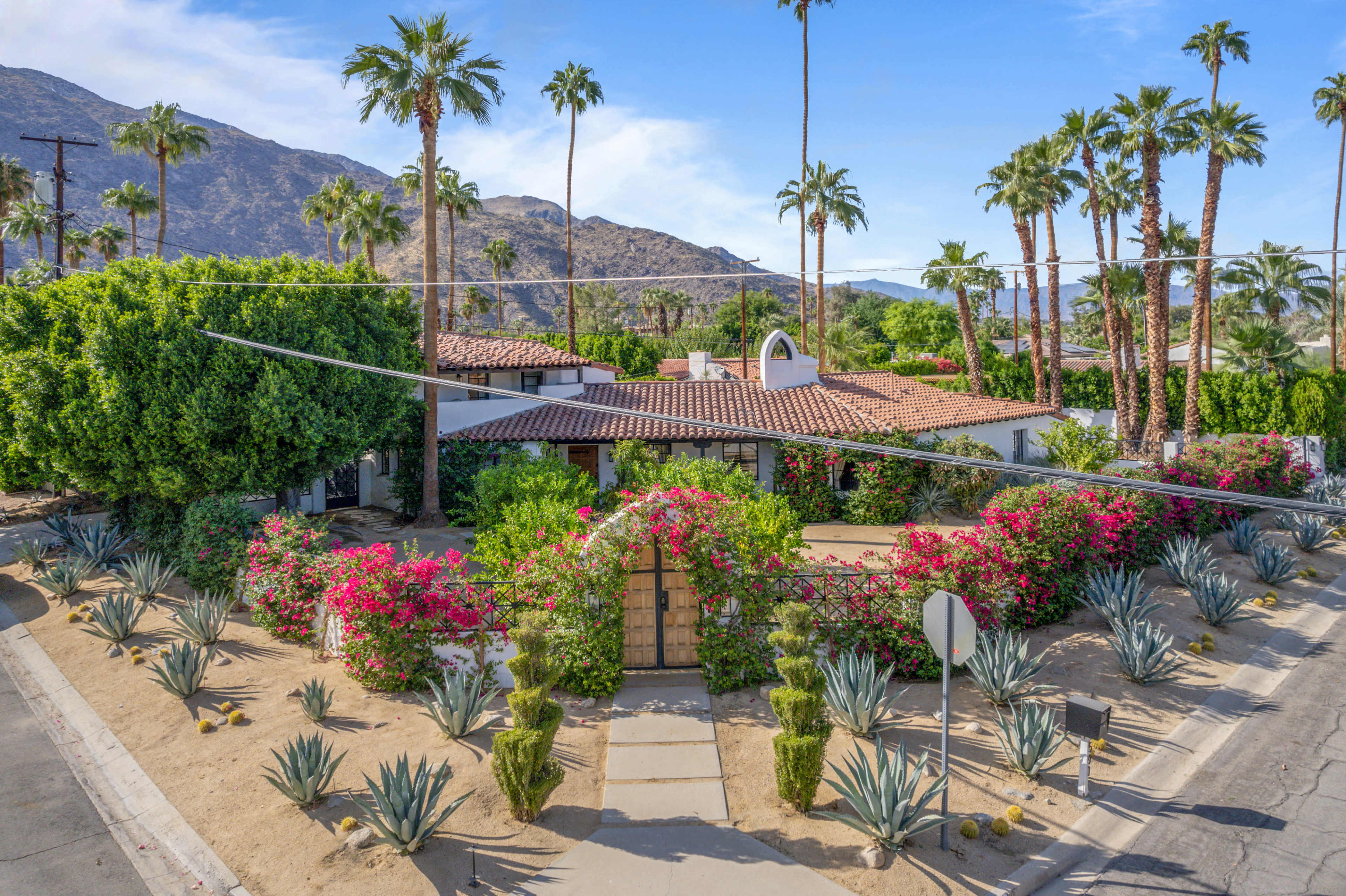 A single-story house surrounded by palm trees and flowering shrubs, with a pathway leading to a wooden gate and mountains in the background.