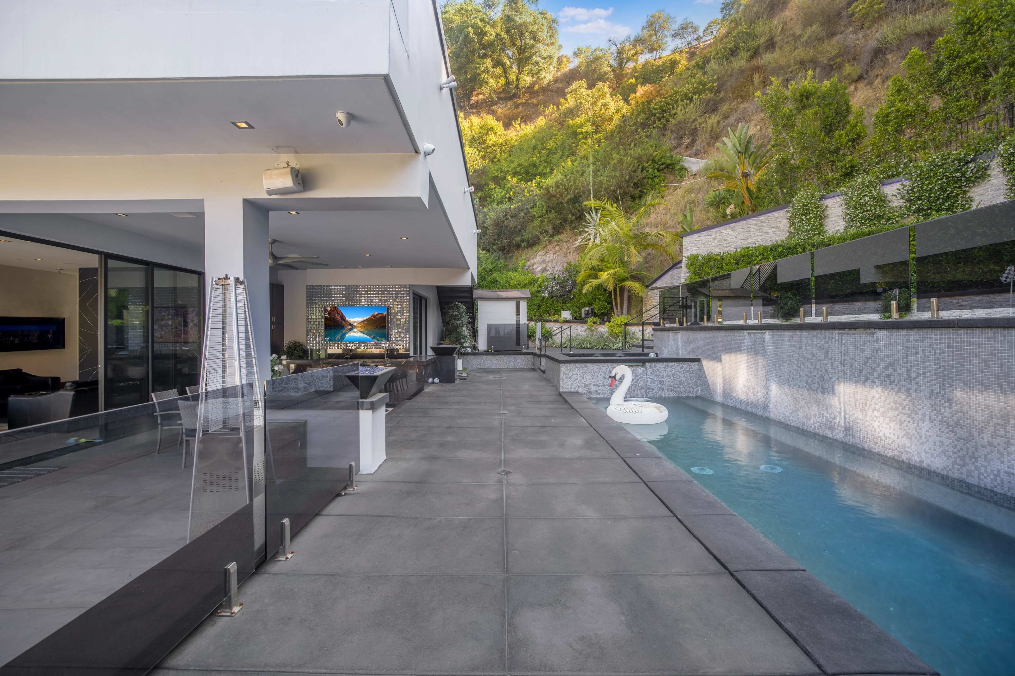 A modern patio features a swimming pool with a swan float, surrounded by greenery and a hillside.