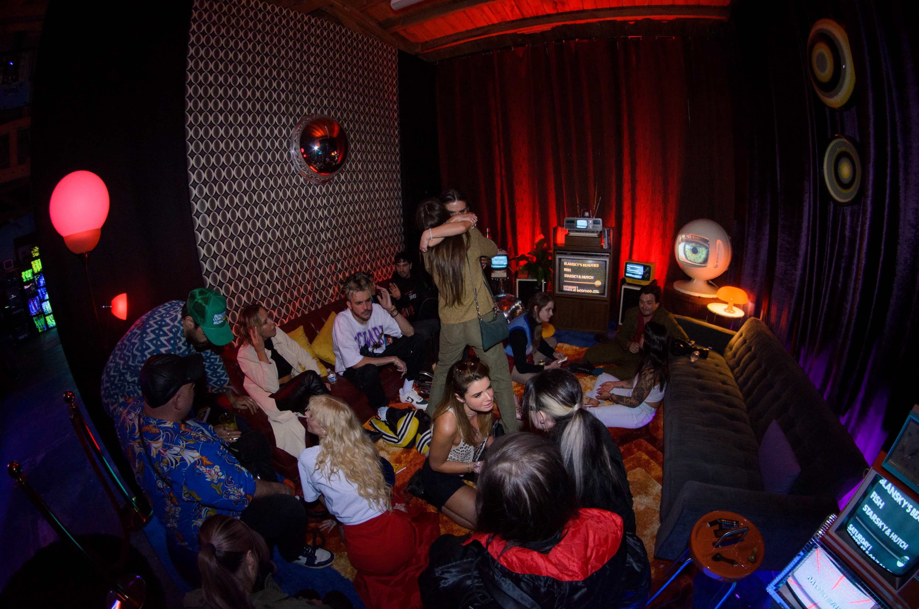 A group of people is gathered in a cozy room with retro decor, chatting and sitting on a large couch while some are embracing.