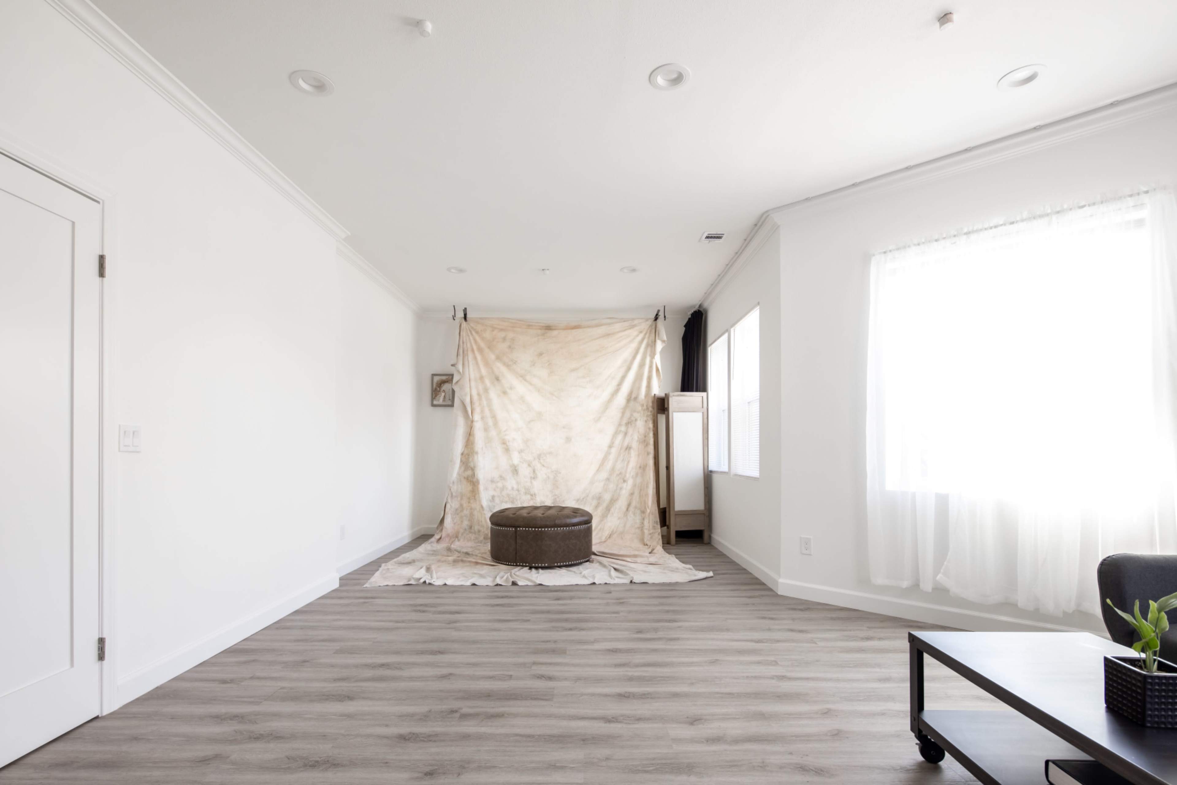 A spacious room with light-colored walls and a wooden floor, featuring a fabric backdrop and a round ottoman in the center.