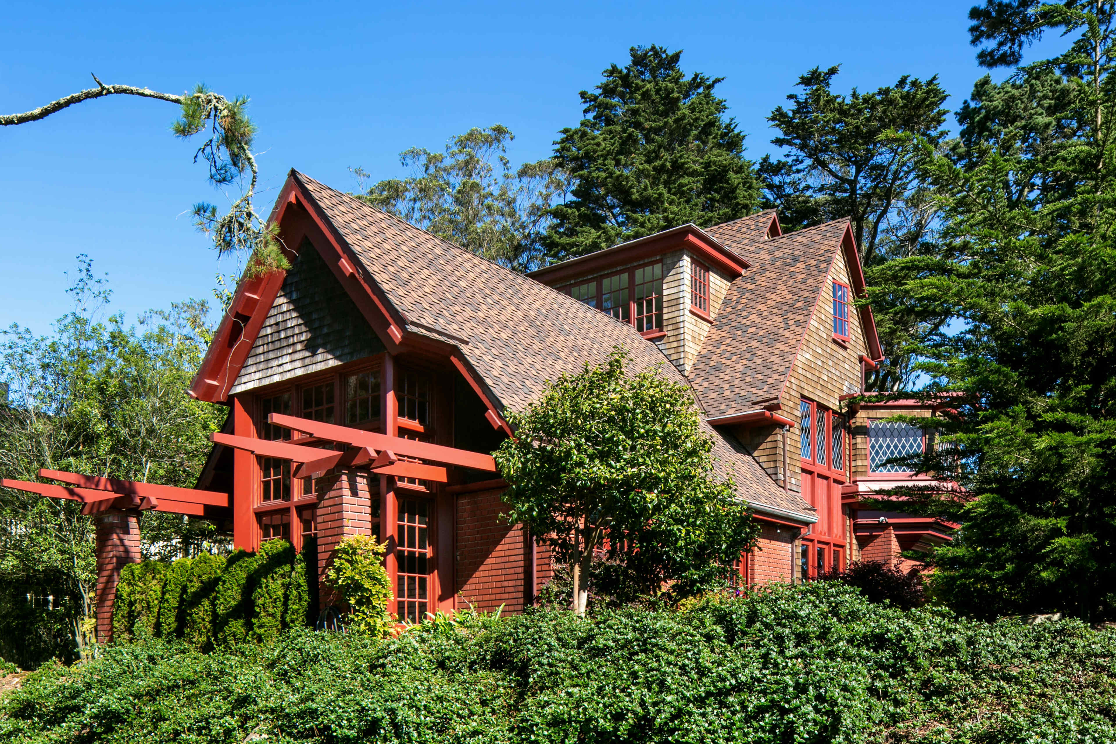 A large, two-story house with a unique architectural style, featuring a steeply pitched roof and red-painted wooden accents, surrounded by greenery.