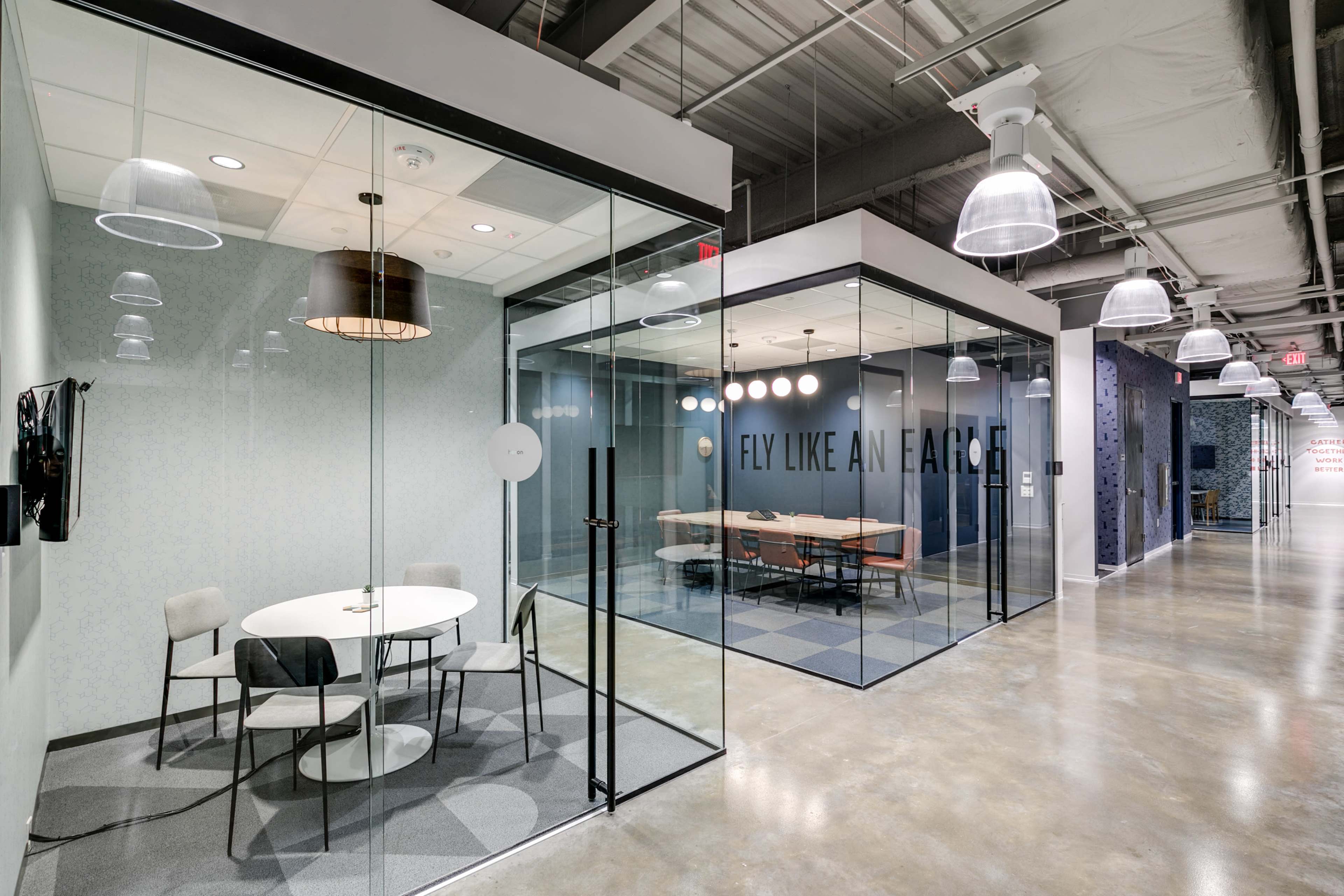 The image shows a modern office space with glass-walled meeting rooms, a circular table, and a motivational slogan on the wall.