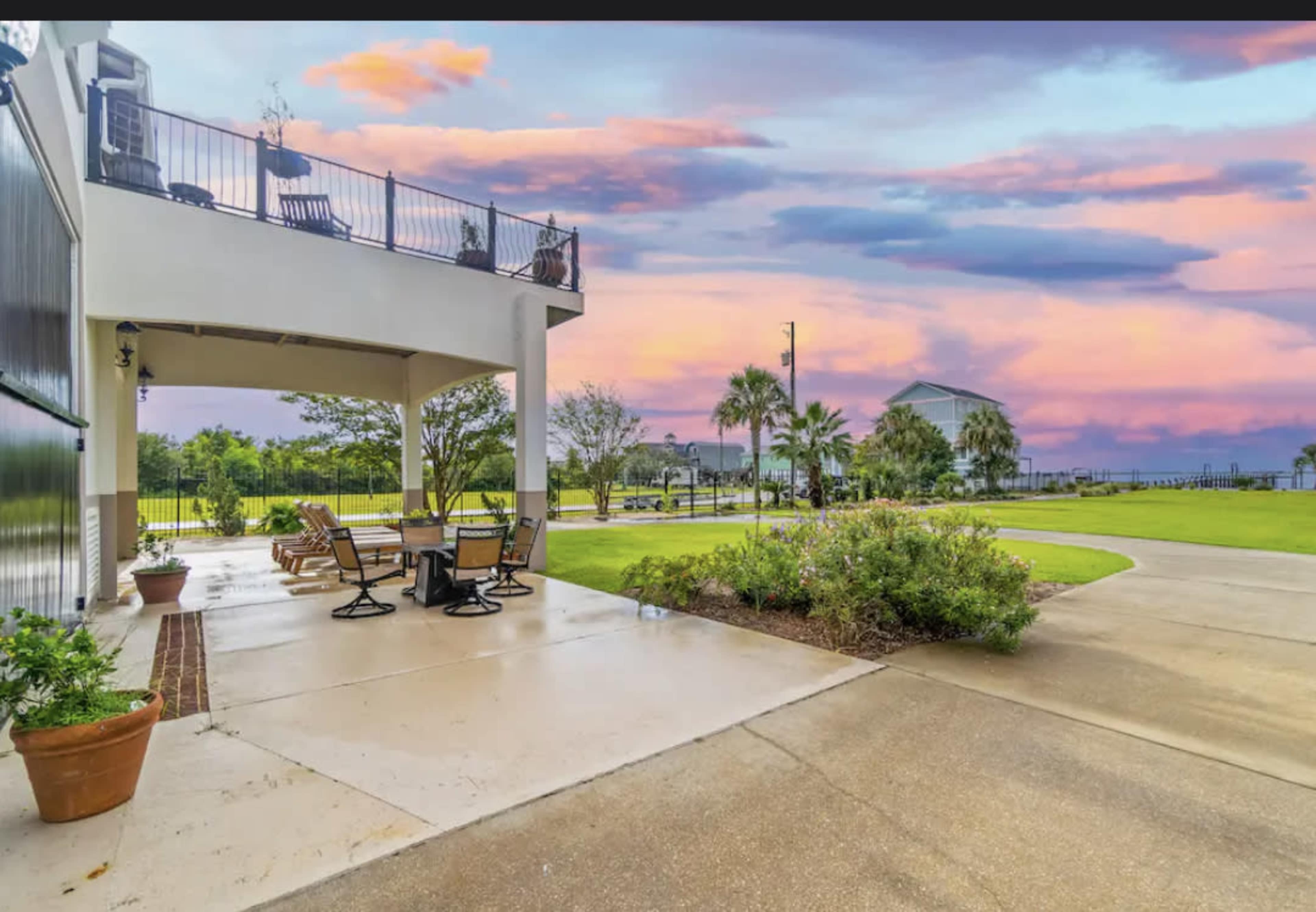 A patio area with outdoor furniture, bordered by lush greenery, under a colorful sunset sky.