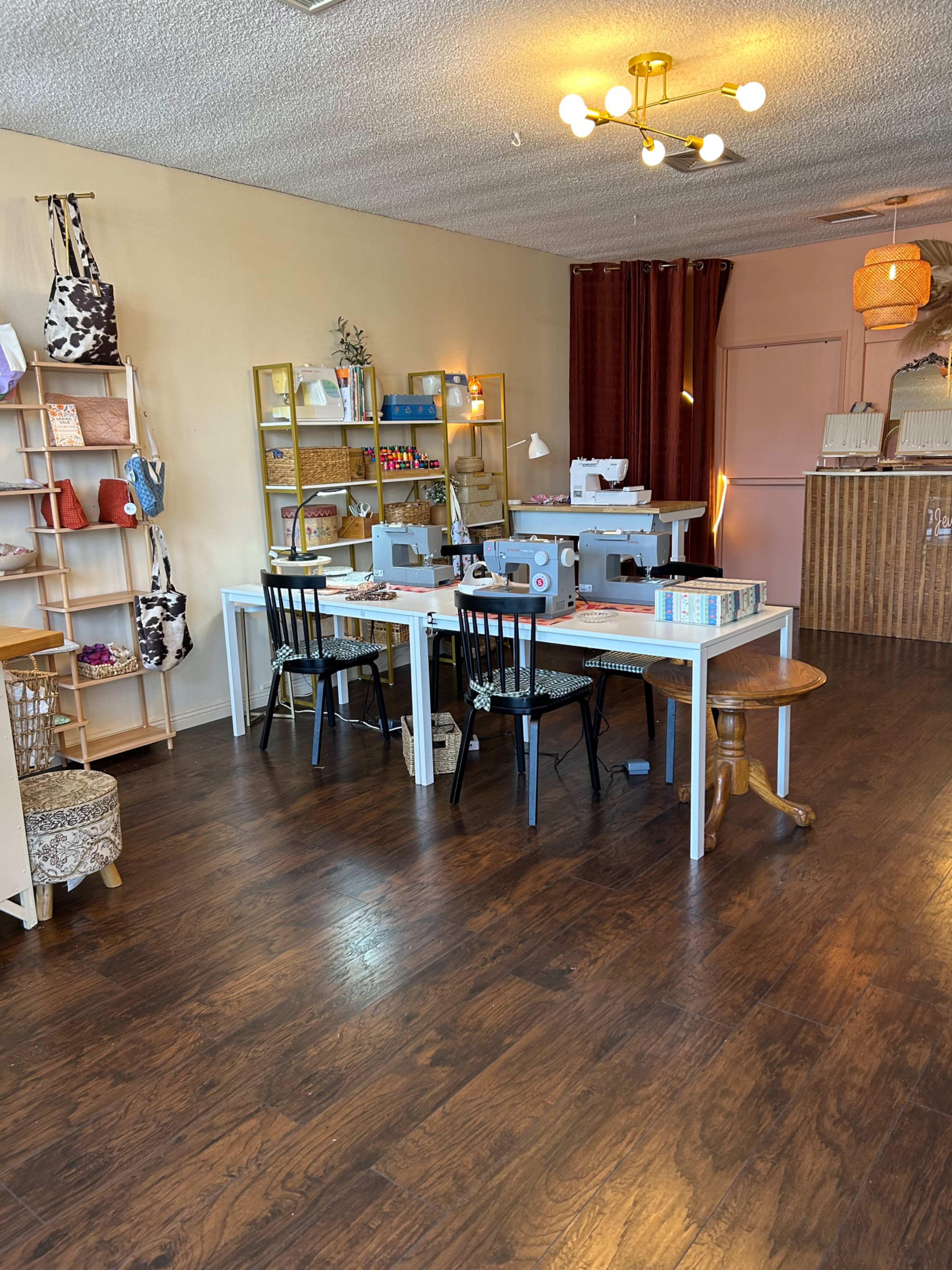 The image shows a craft studio with sewing machines on a central table, surrounded by chairs, and shelves displaying various crafting supplies.