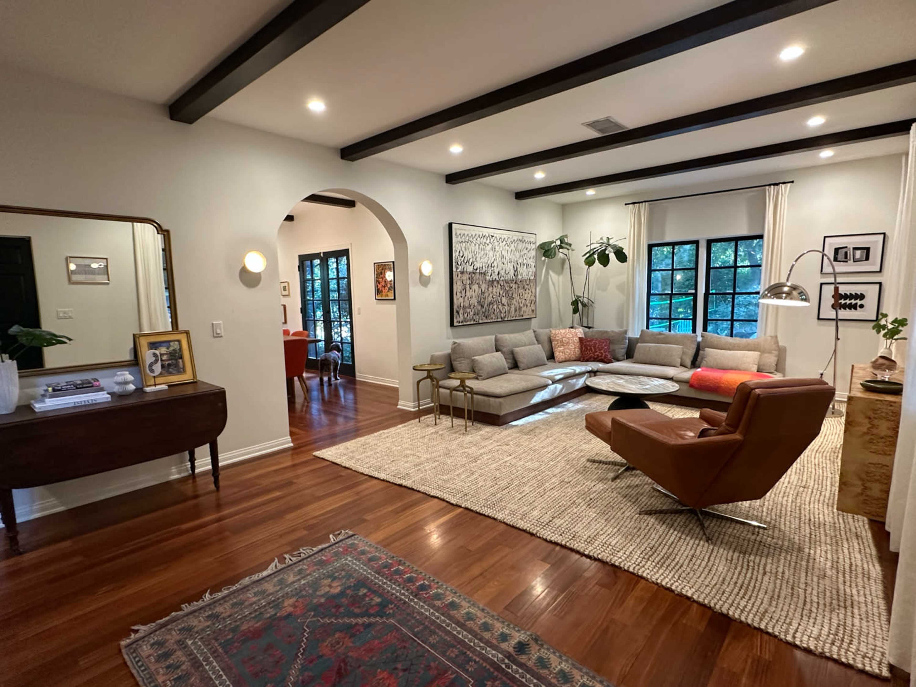 A living room featuring a large seating arrangement, a coffee table, a decorative rug, and various plants, all set against a backdrop of exposed ceiling beams and large windows.