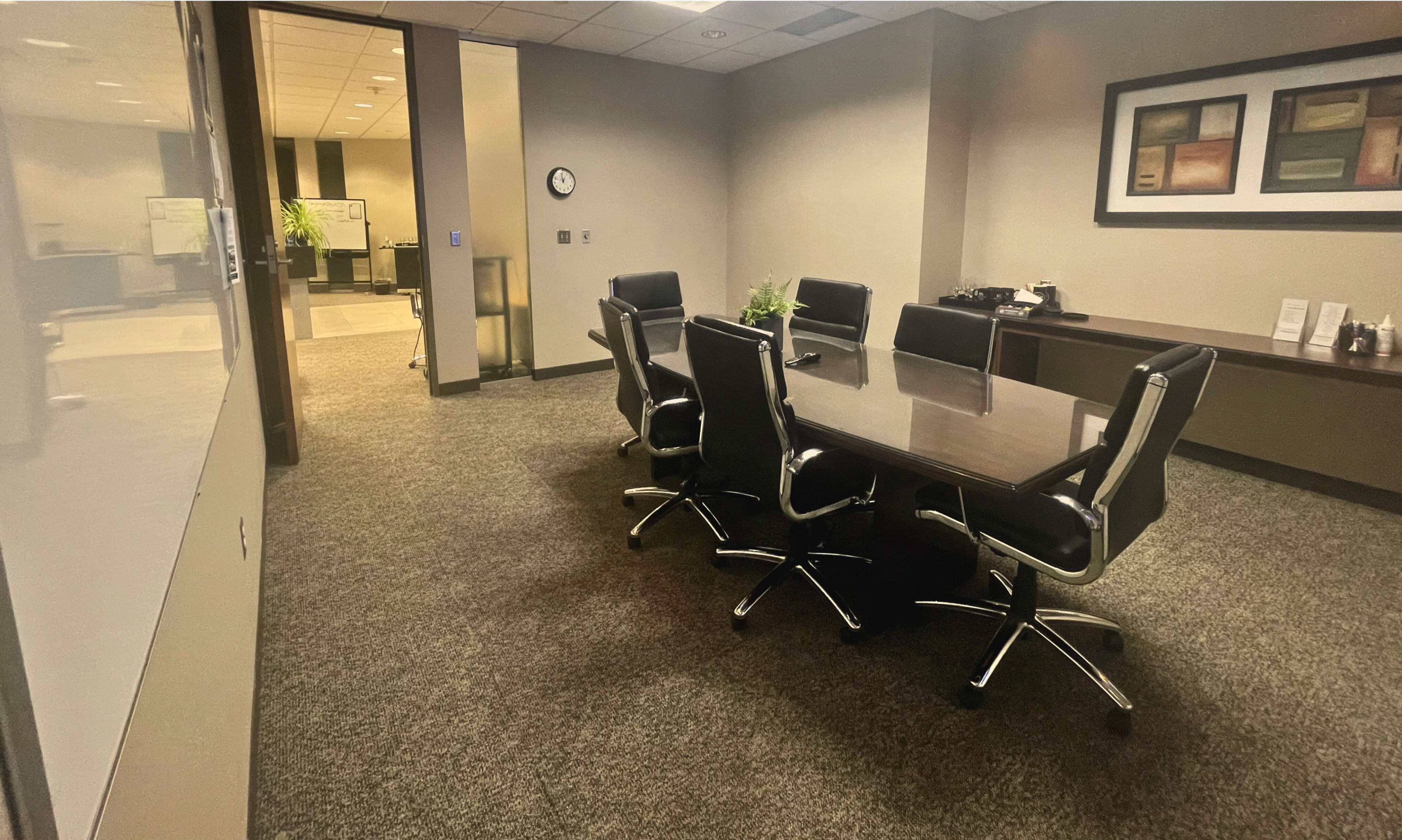The image shows a conference room with a large table surrounded by six black chairs, and a wall with framed artwork.