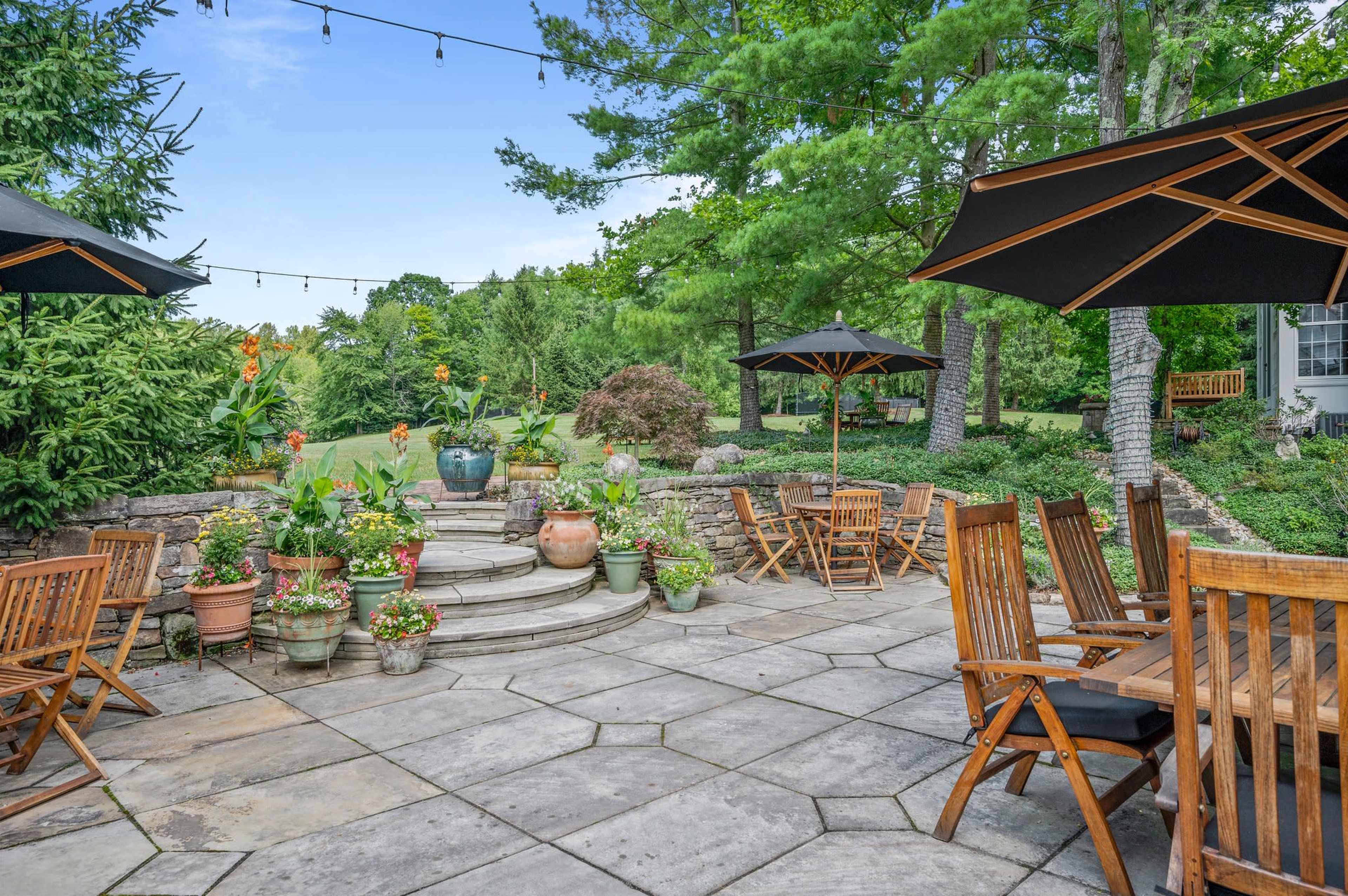 The image shows a landscaped patio area with wooden furniture, potted plants, and umbrellas, surrounded by greenery and a stone pathway.