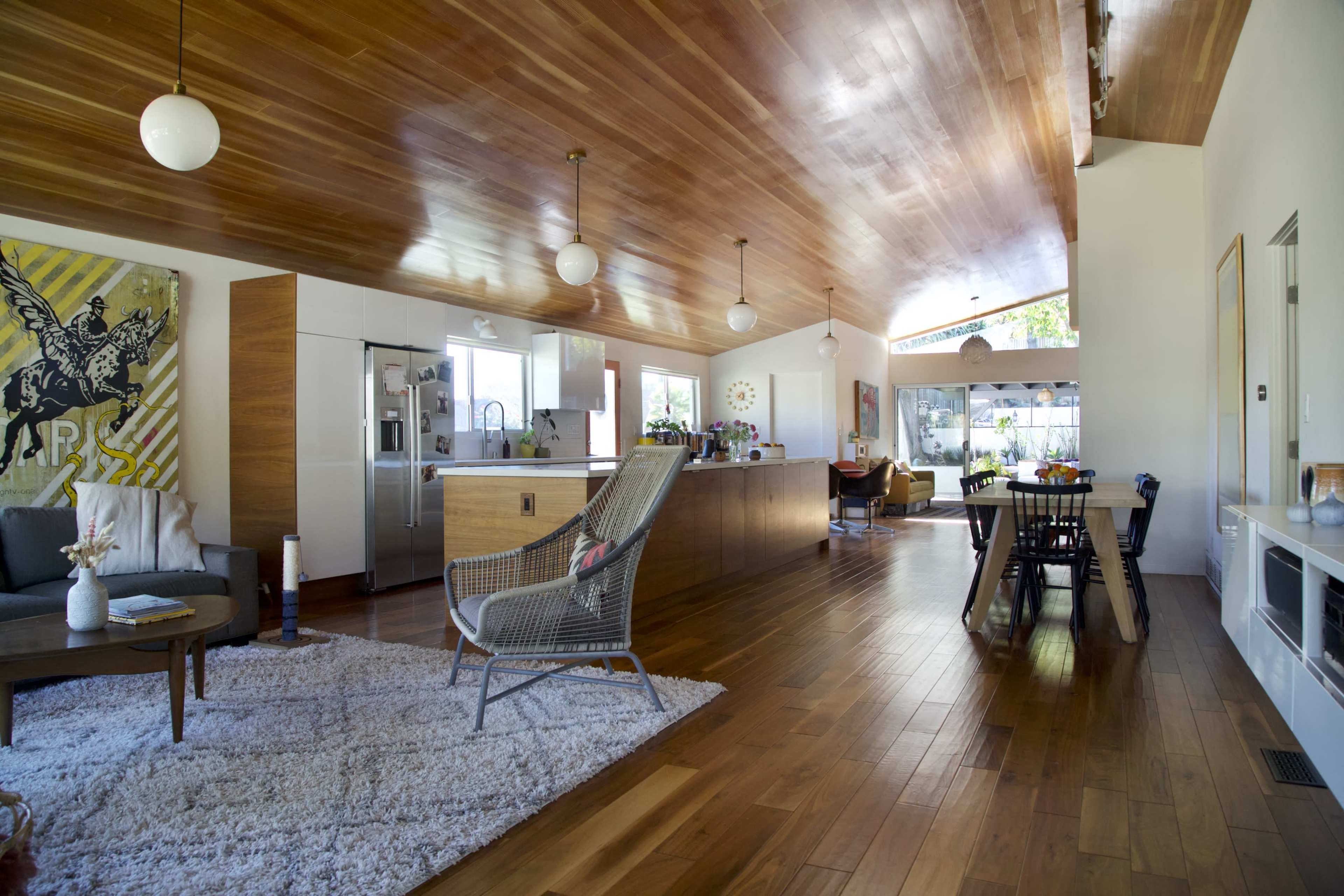 A spacious, modern living area featuring a wooden ceiling, a kitchen with an island, and a dining area adjacent to large windows.