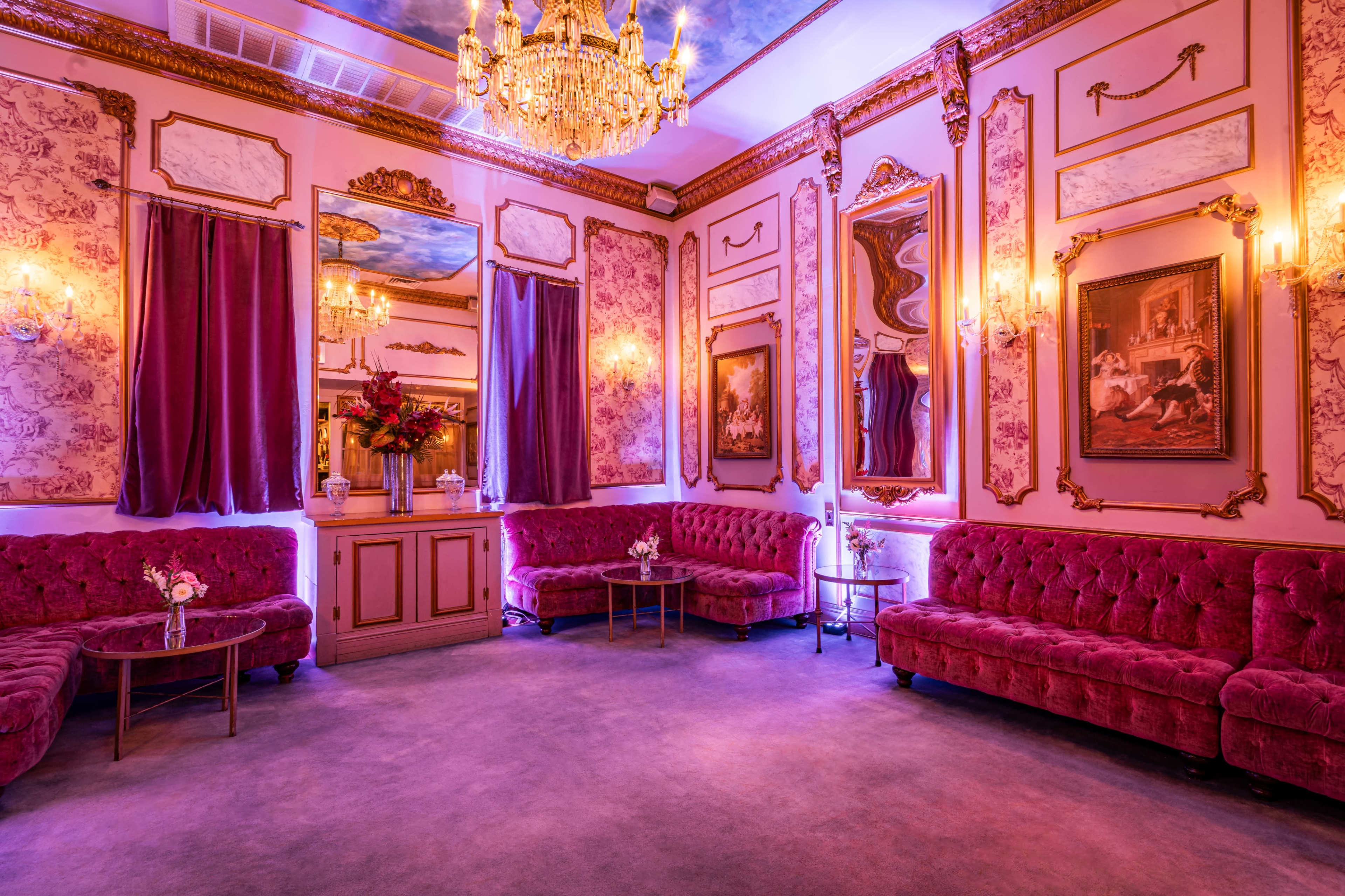 The image shows an elegantly decorated room featuring plush purple seating, ornate mirrors, and a chandelier.