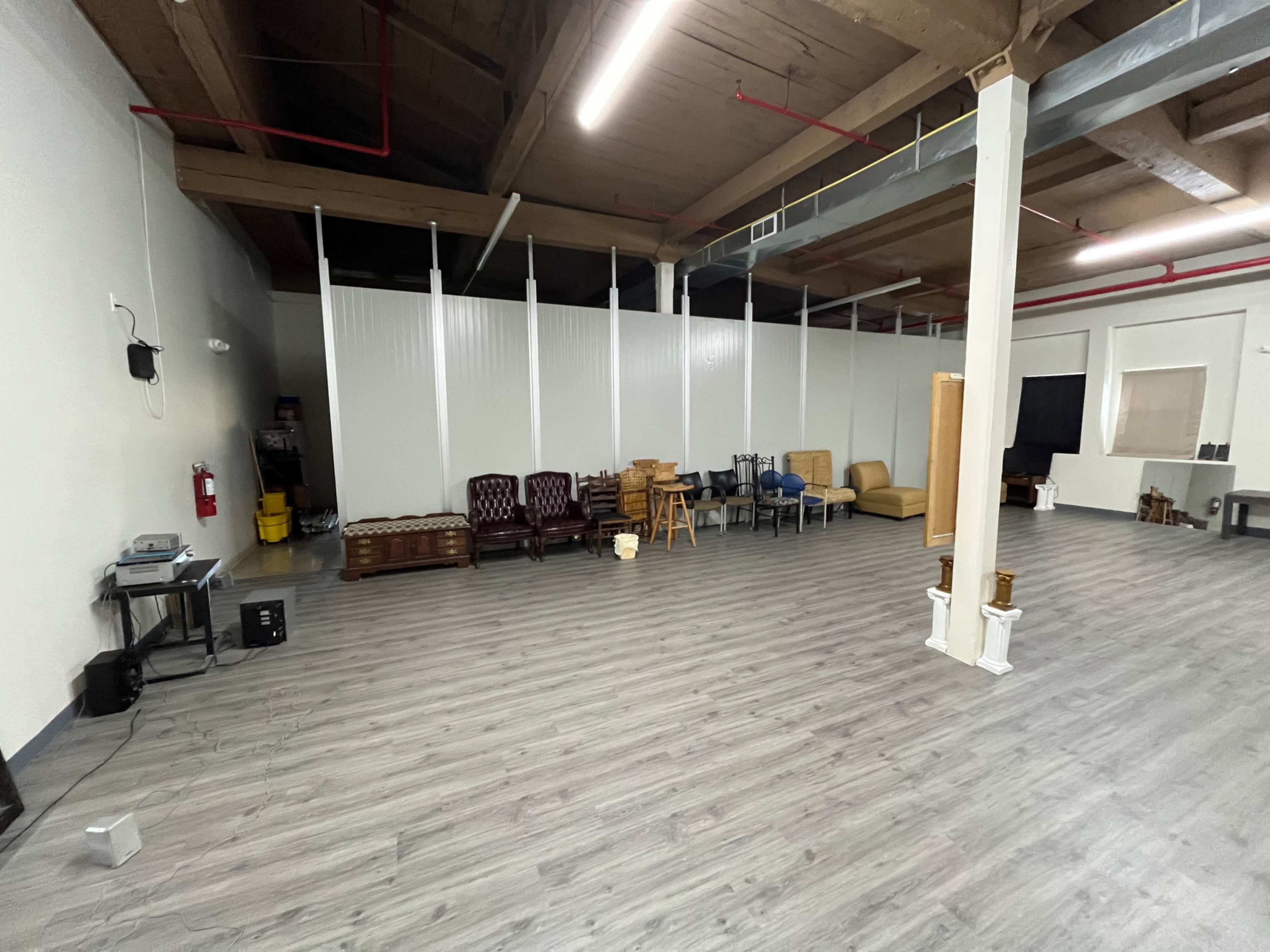 The image shows a spacious interior room with wooden flooring, white walls, and various seating furniture arranged against a partition.