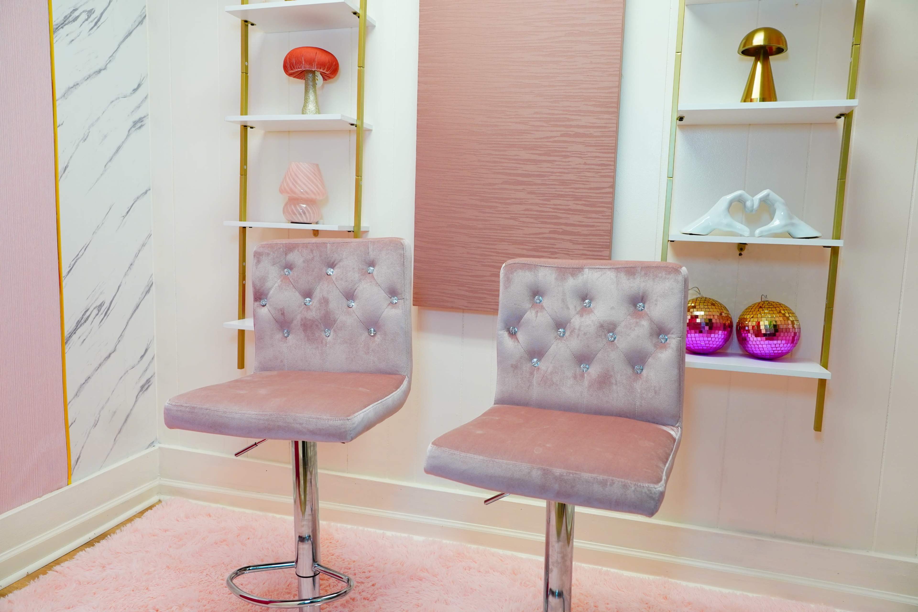 The image features two pink velvet bar chairs in front of a stylish background with decorative shelves displaying various colorful objects.