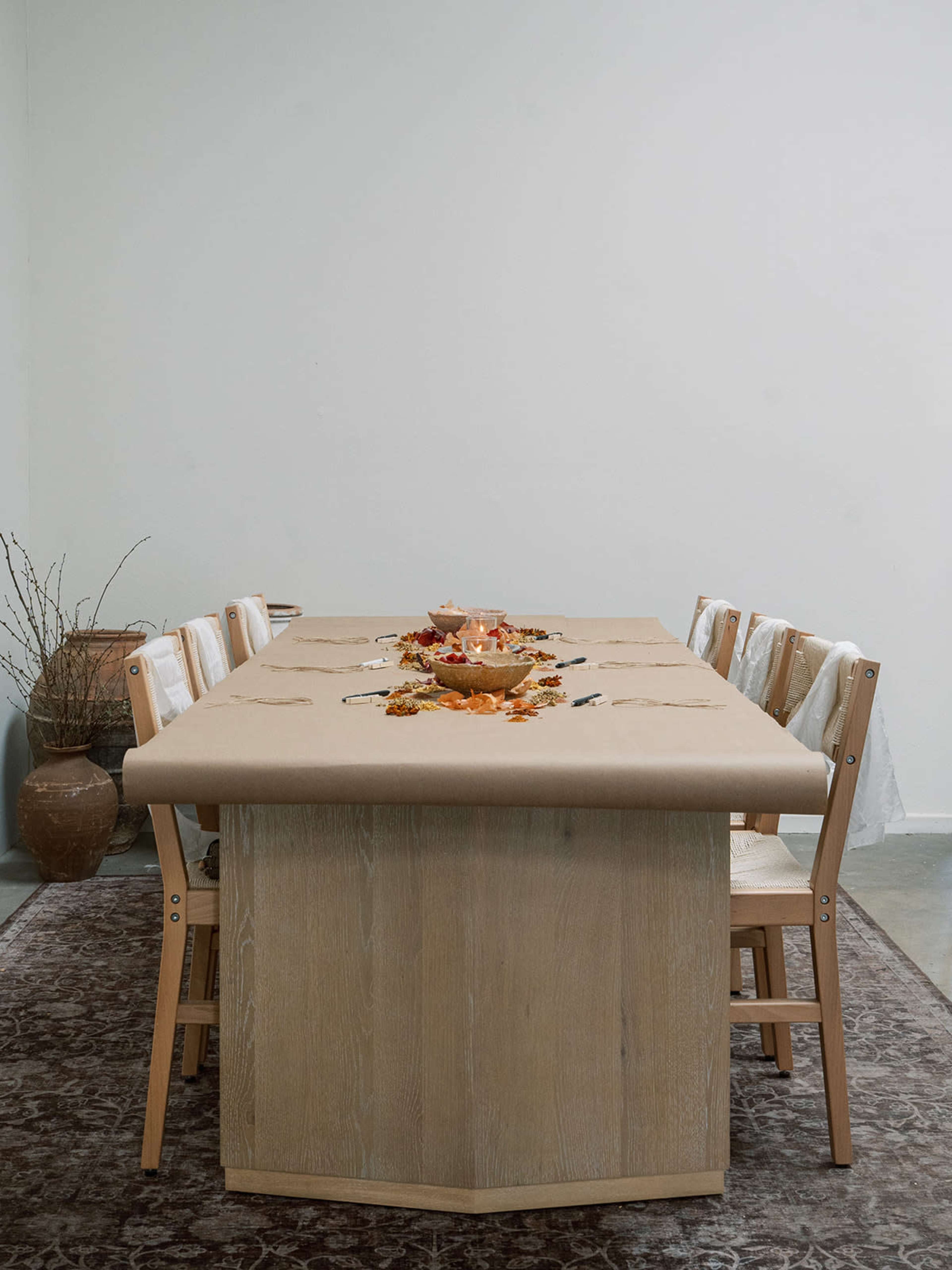 A wooden dining table is set with a brown paper tablecloth and a bowl of decorative fruits, surrounded by wooden chairs in a neutral-toned room.