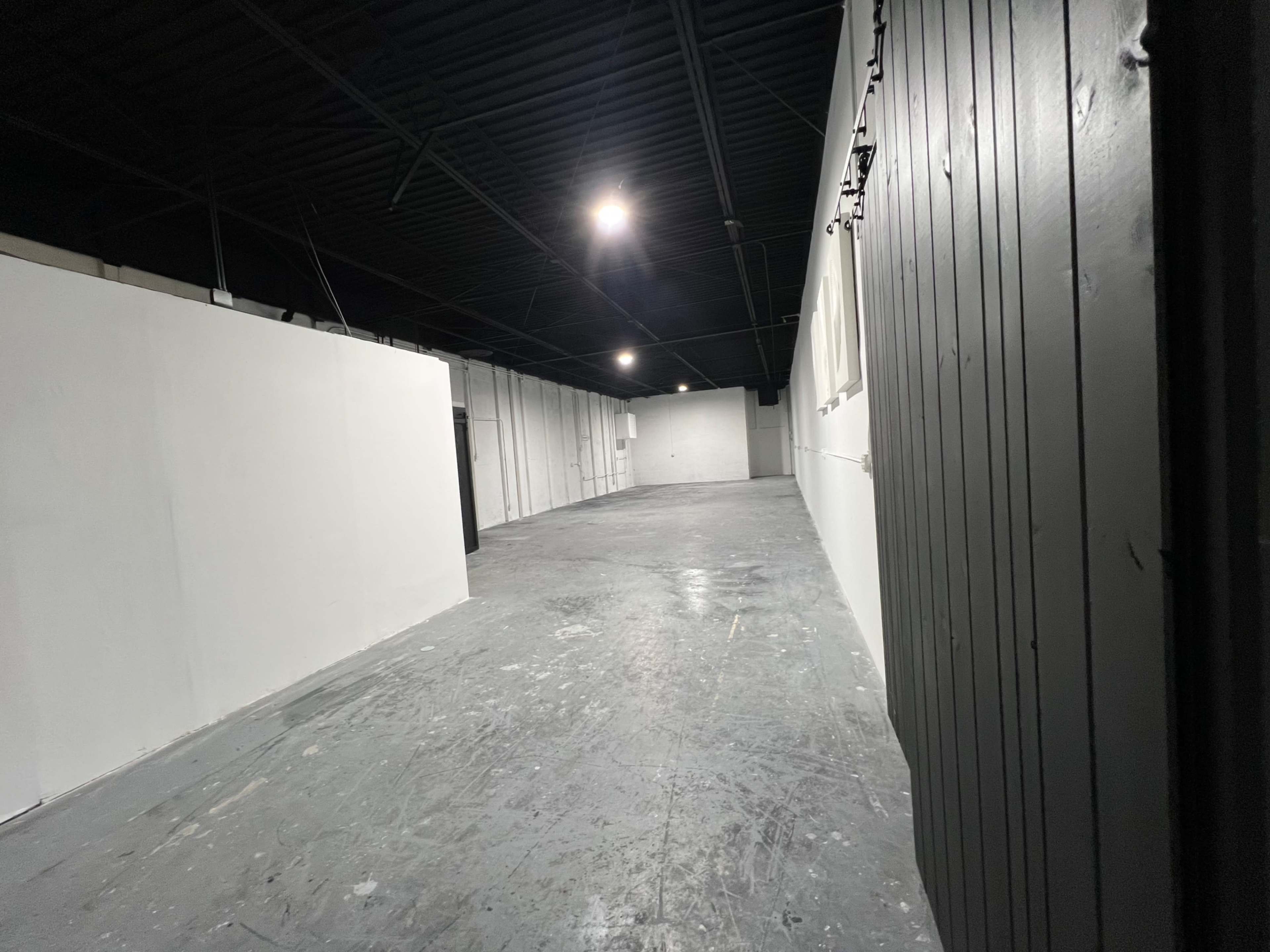 The image shows an empty, spacious interior with gray concrete flooring and white walls, illuminated by overhead lights.