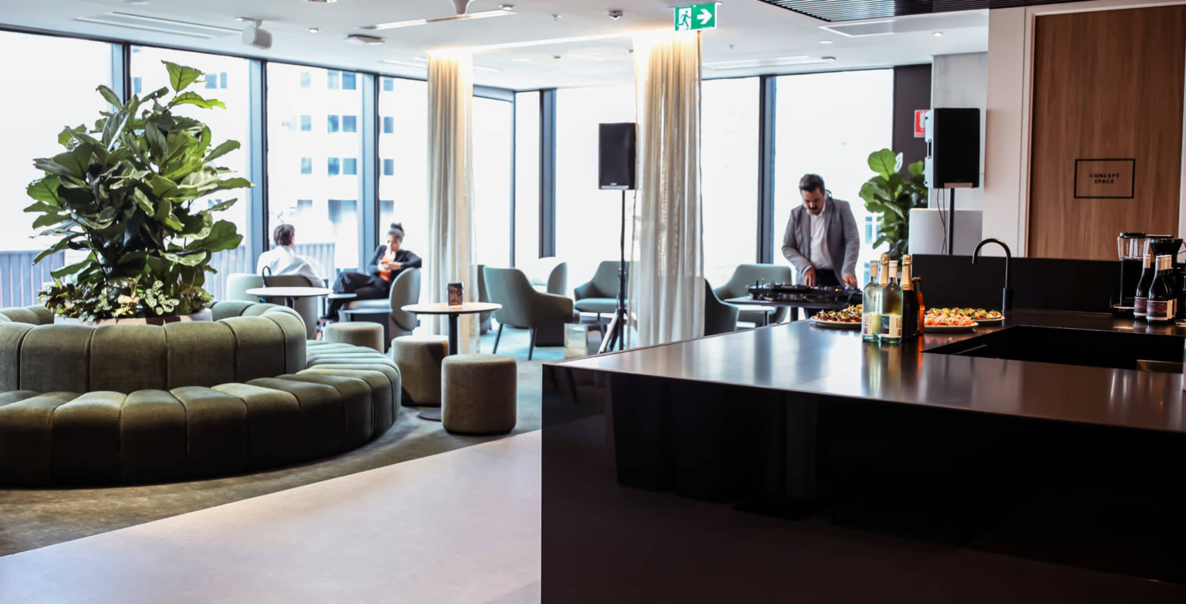 A modern lounge area features a curved green seating arrangement, tables, and a man playing music while others converse in the background.