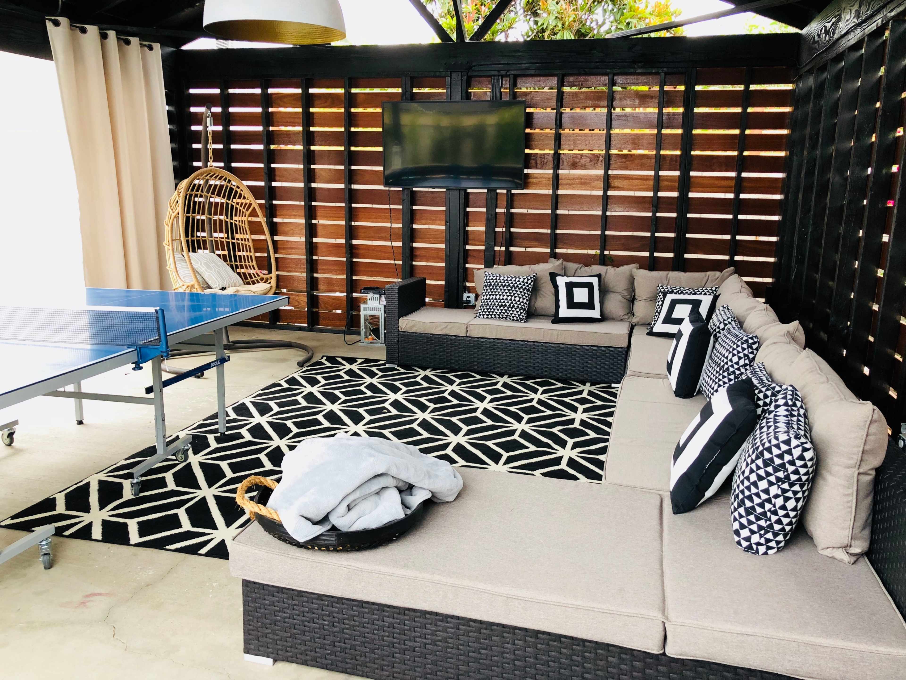 A covered outdoor seating area with a sectional sofa, decorative pillows, a television on the wall, a ping pong table, and a patterned rug.