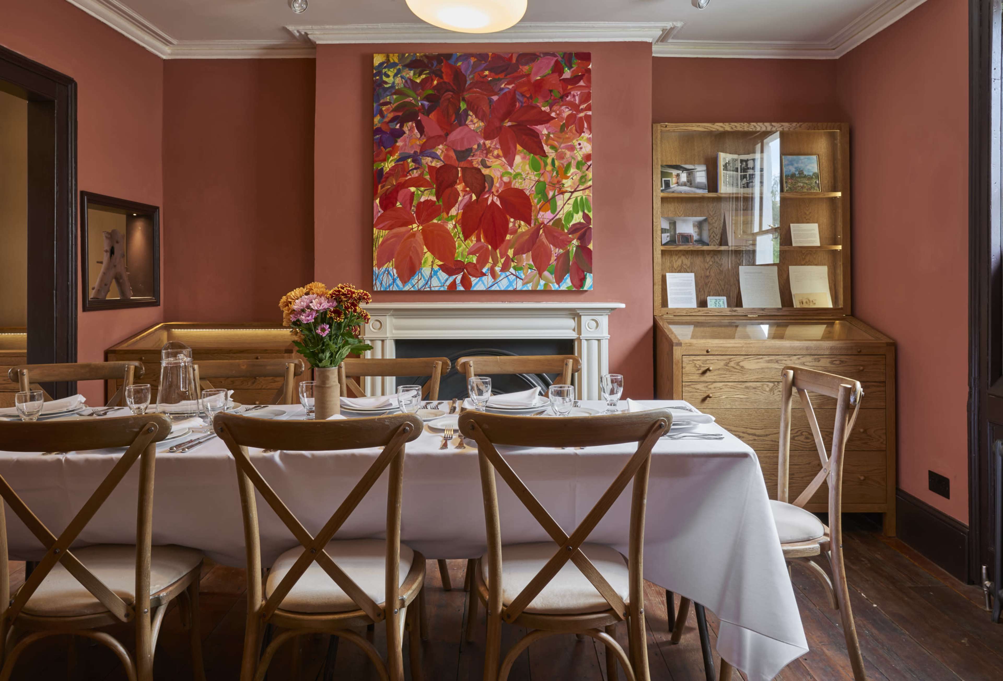 A dining room features a large wooden table set with white tableware, surrounded by chairs, against a backdrop of a colorful wall painting and a wooden cabinet.