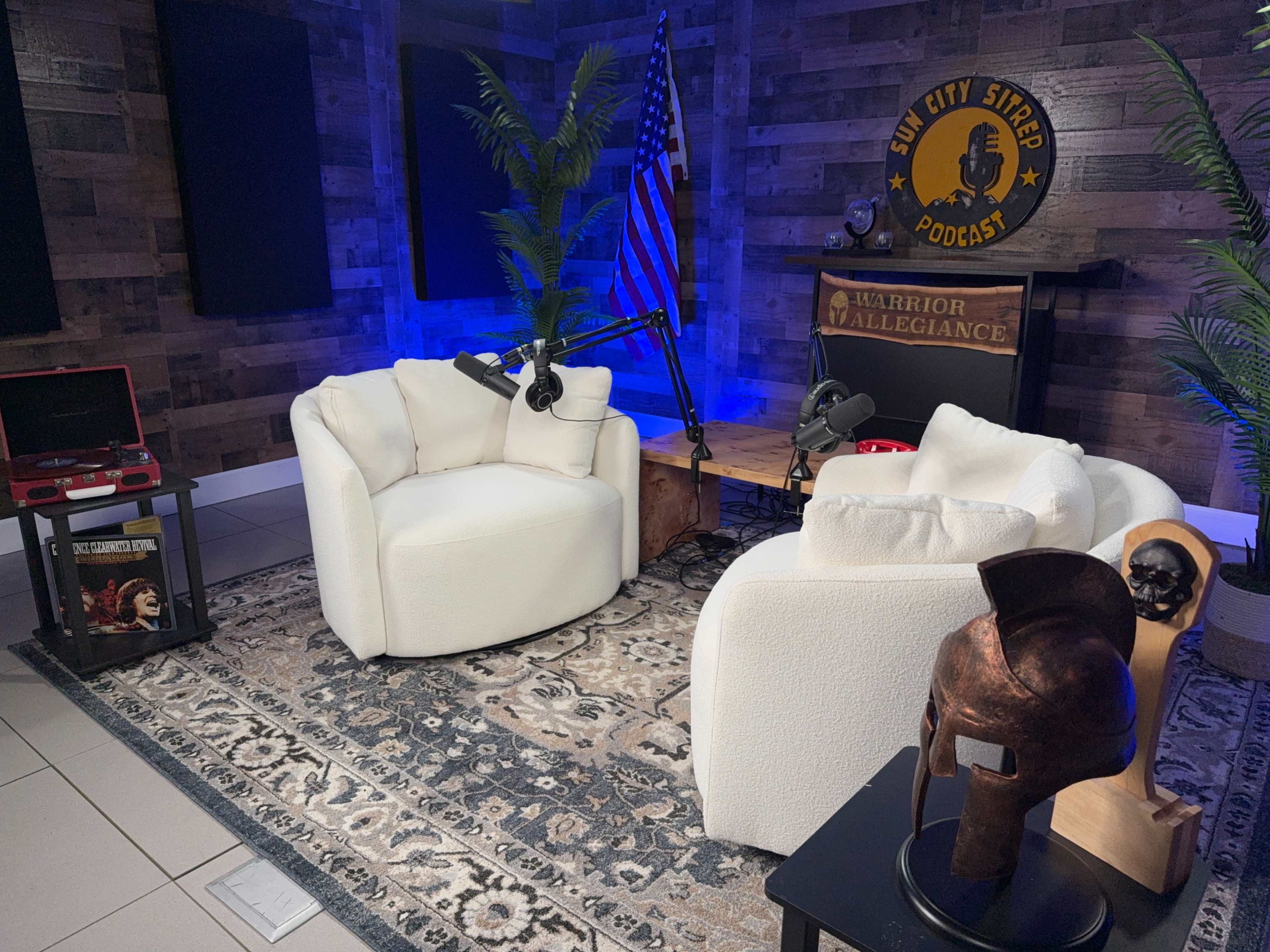 The image shows a podcast set featuring two white chairs, a wooden table, a microphone setup, and decorative elements like a flag and a statue.