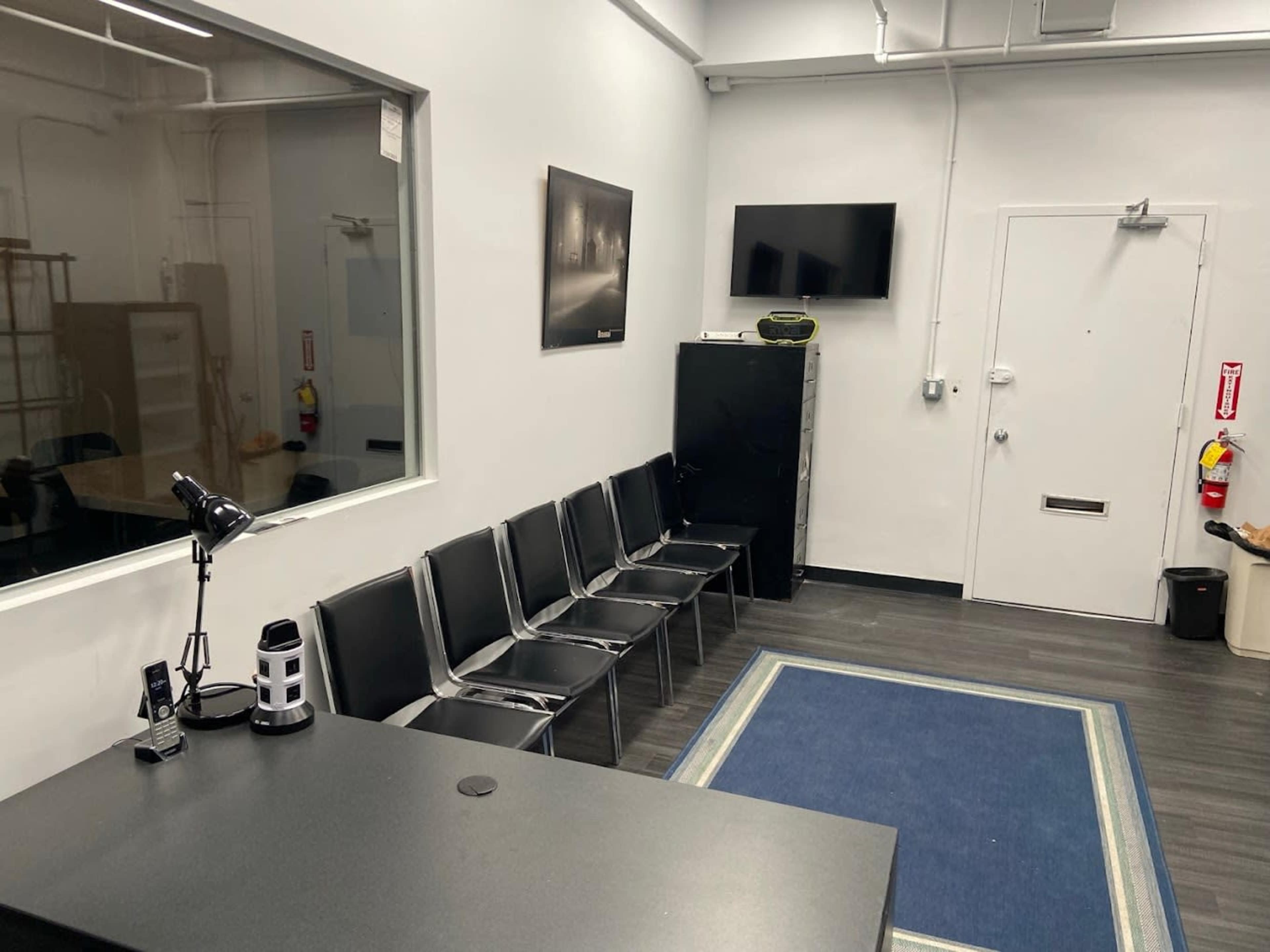 The image shows a waiting area with black chairs arranged along a wall, a desk with a lamp and a small device, a wall-mounted television, and a door leading to another room.