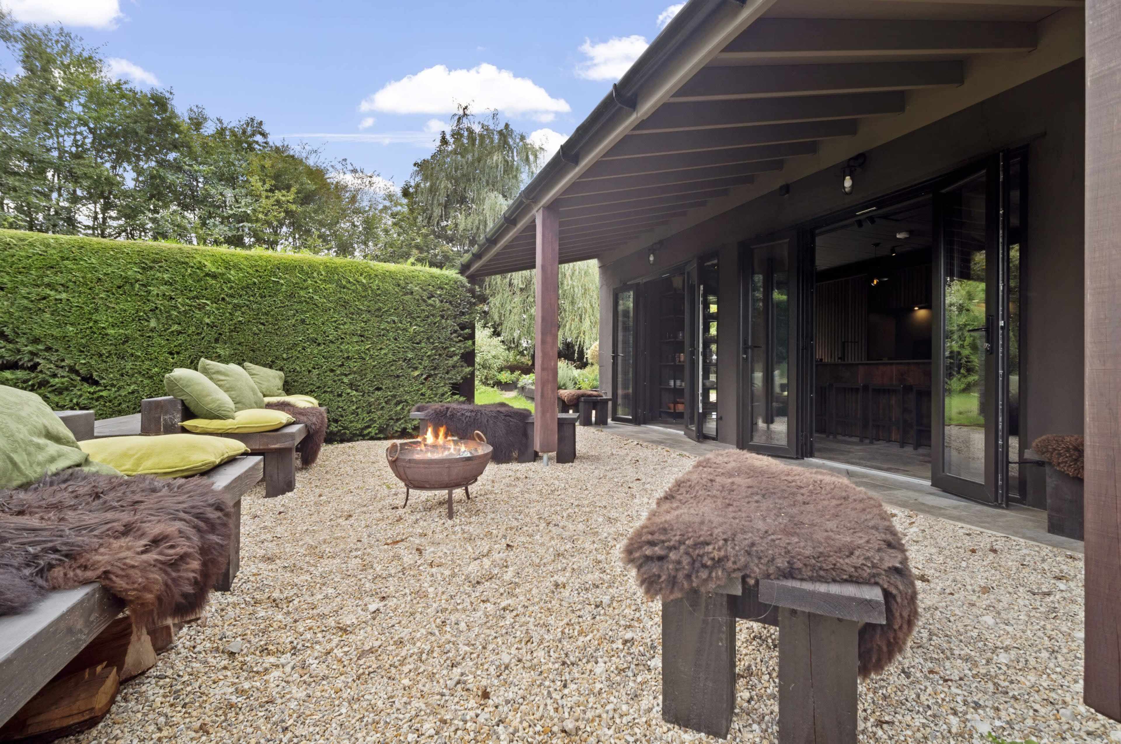 A cozy outdoor seating area features a fire pit surrounded by gravel and benches, with a backdrop of tall hedges and clear blue skies.