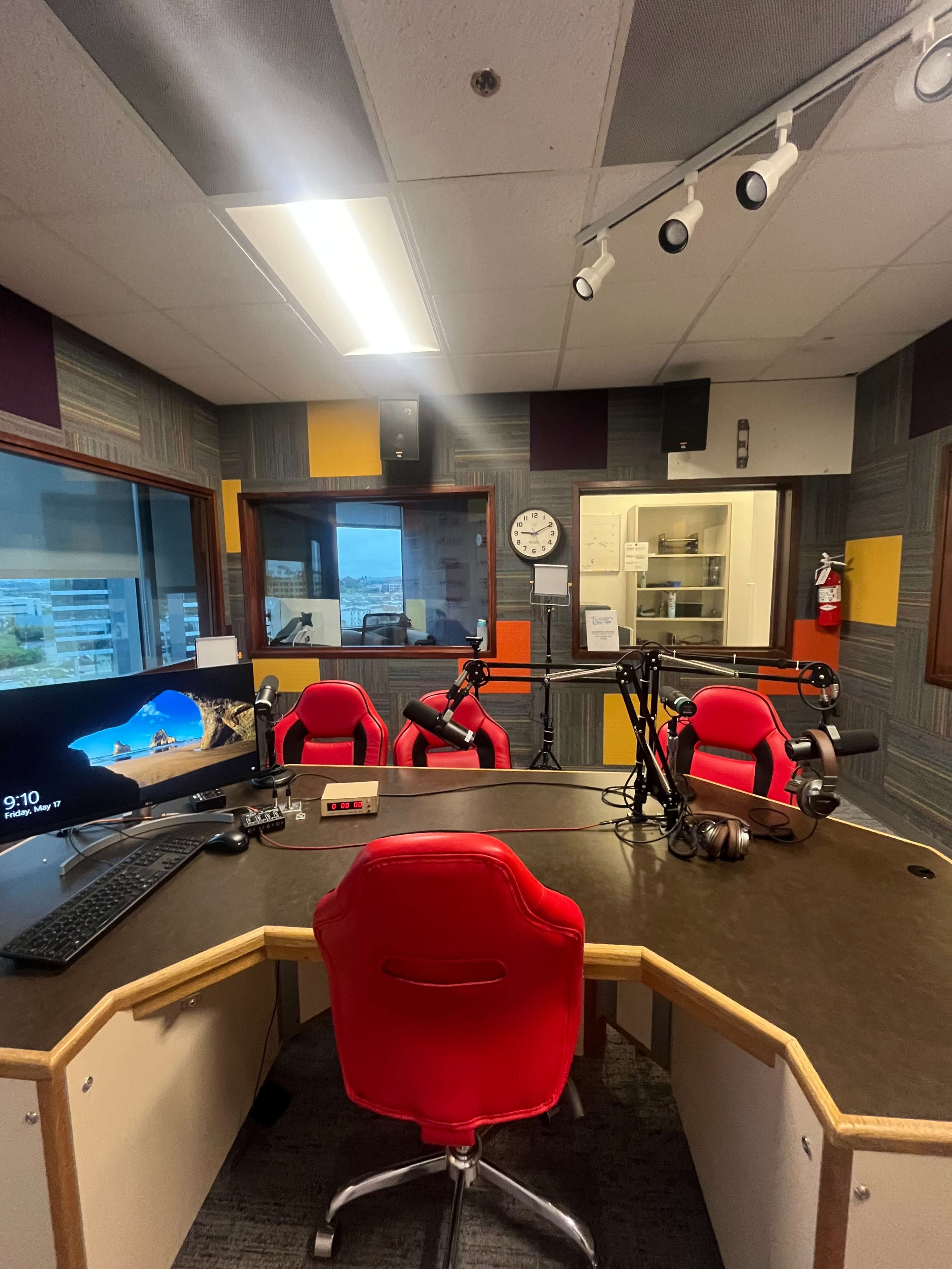 A radio studio featuring a circular desk with red chairs, microphones, and a computer setup, with large windows overlooking the outside.