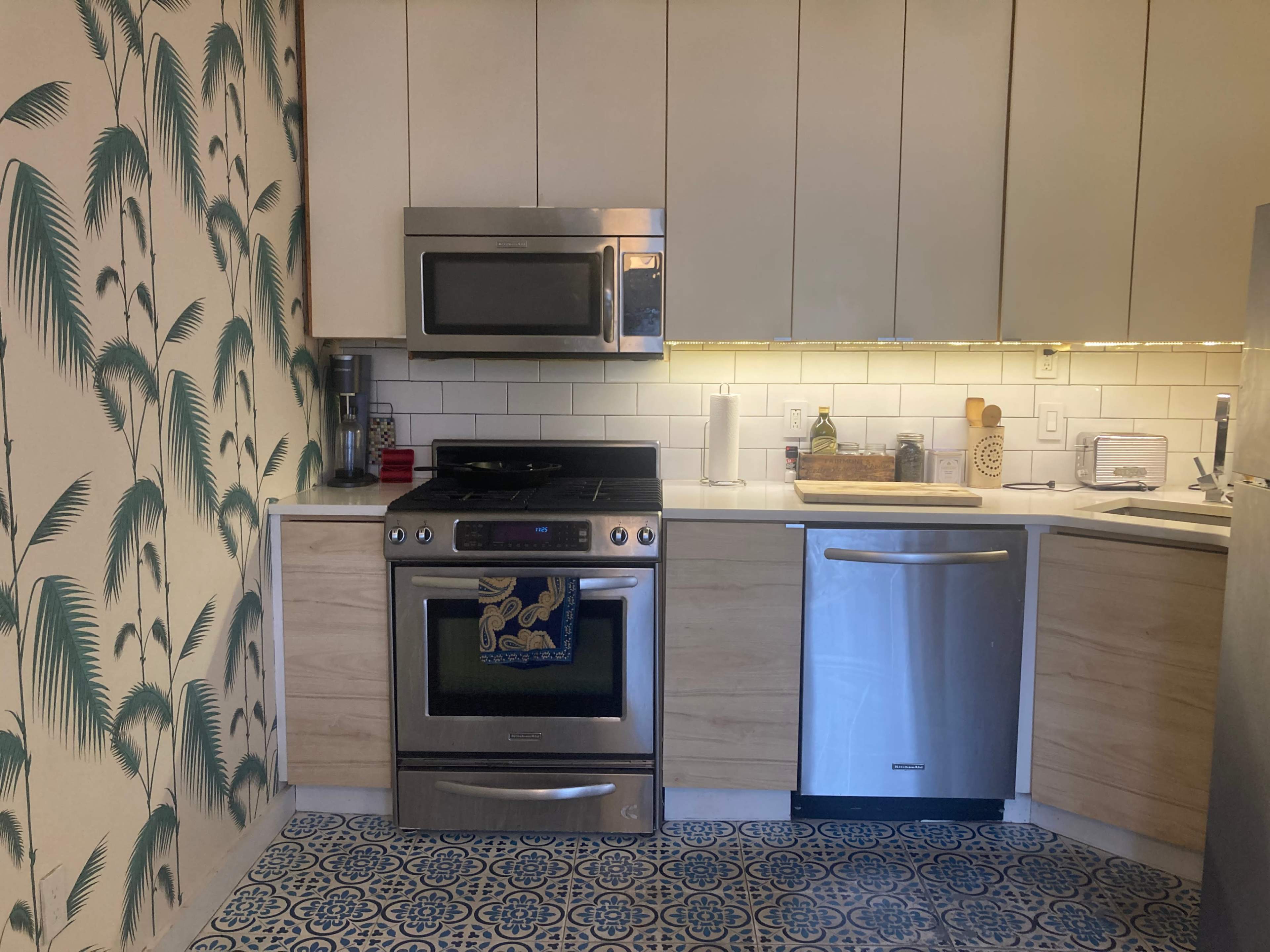 The kitchen features stainless steel appliances, including a stove and microwave, alongside light wood cabinetry and a decorative blue and white tile floor.
