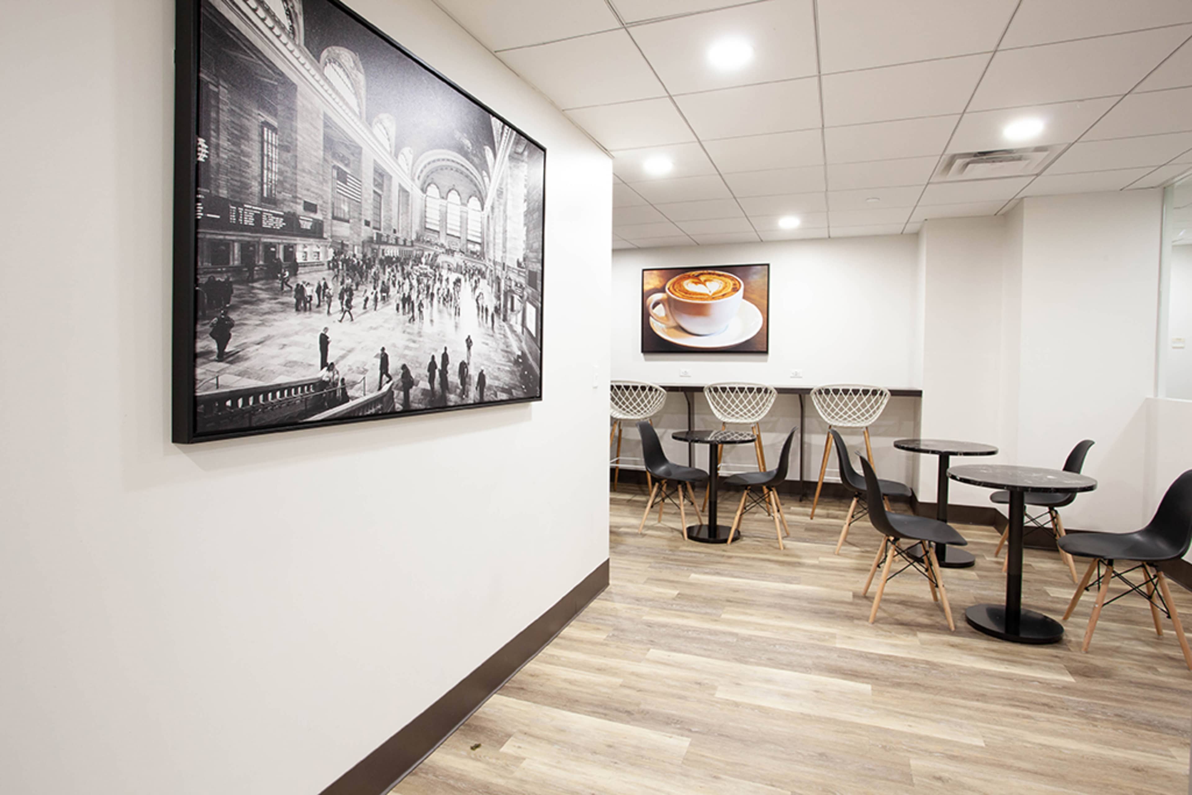 The image shows a modern café interior with a large black-and-white photograph on the wall and several small tables and chairs arranged on a wooden floor.