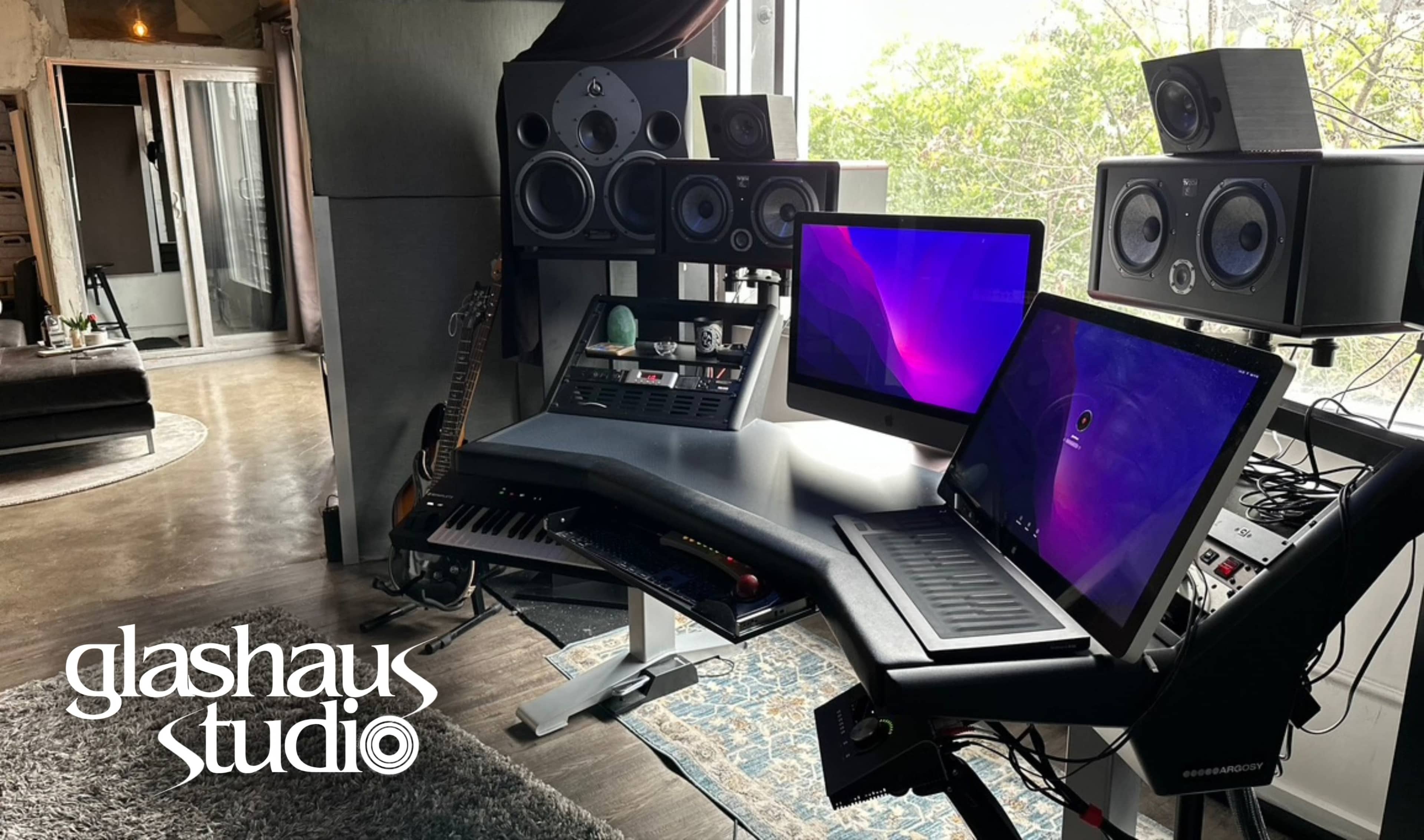 A modern studio workspace featuring two computer monitors, audio speakers, a keyboard, and a guitar against a backdrop of large windows.