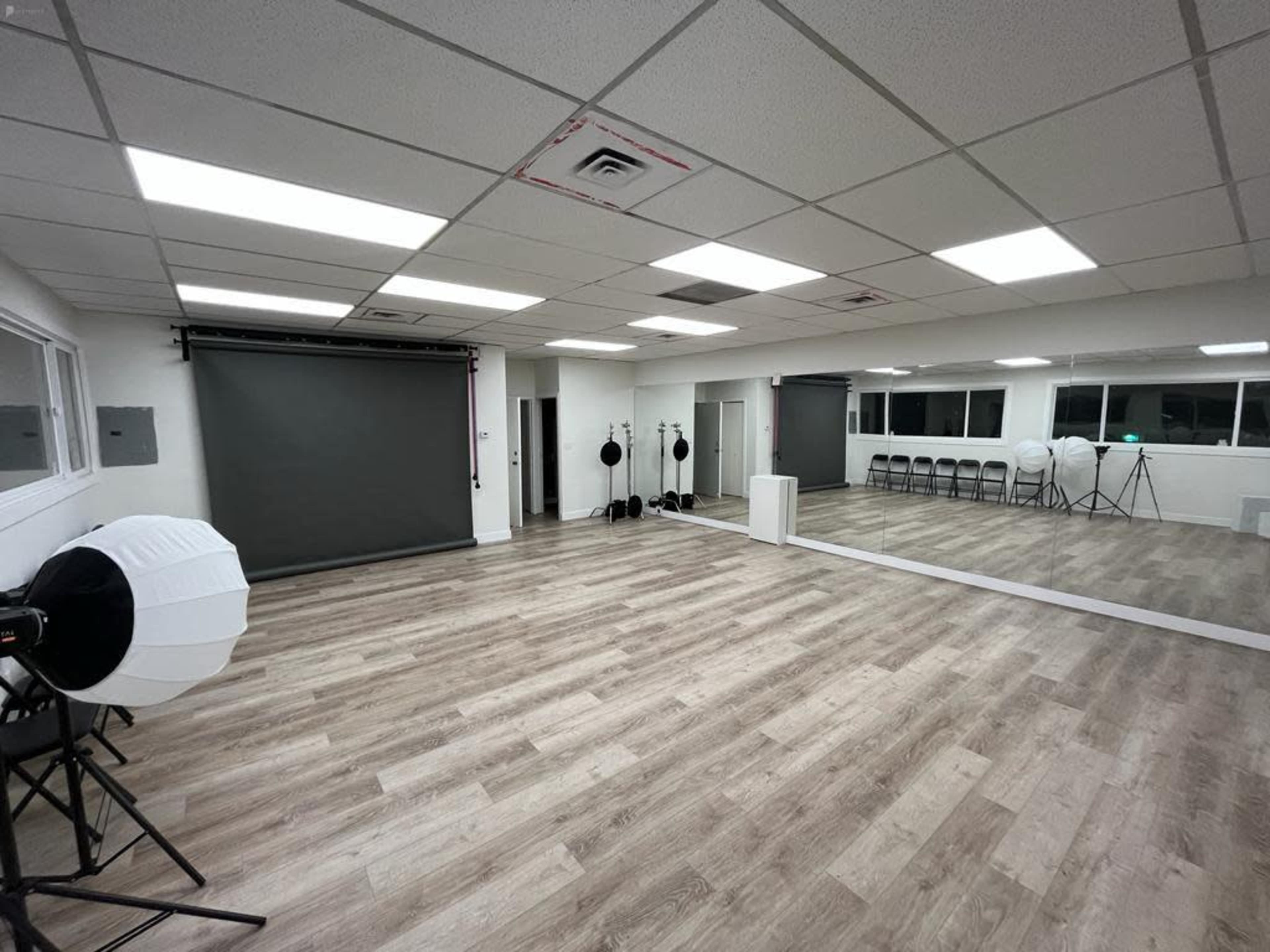 A spacious, well-lit room with a large mirror, a backdrop setup, and several chairs arranged against the wall.