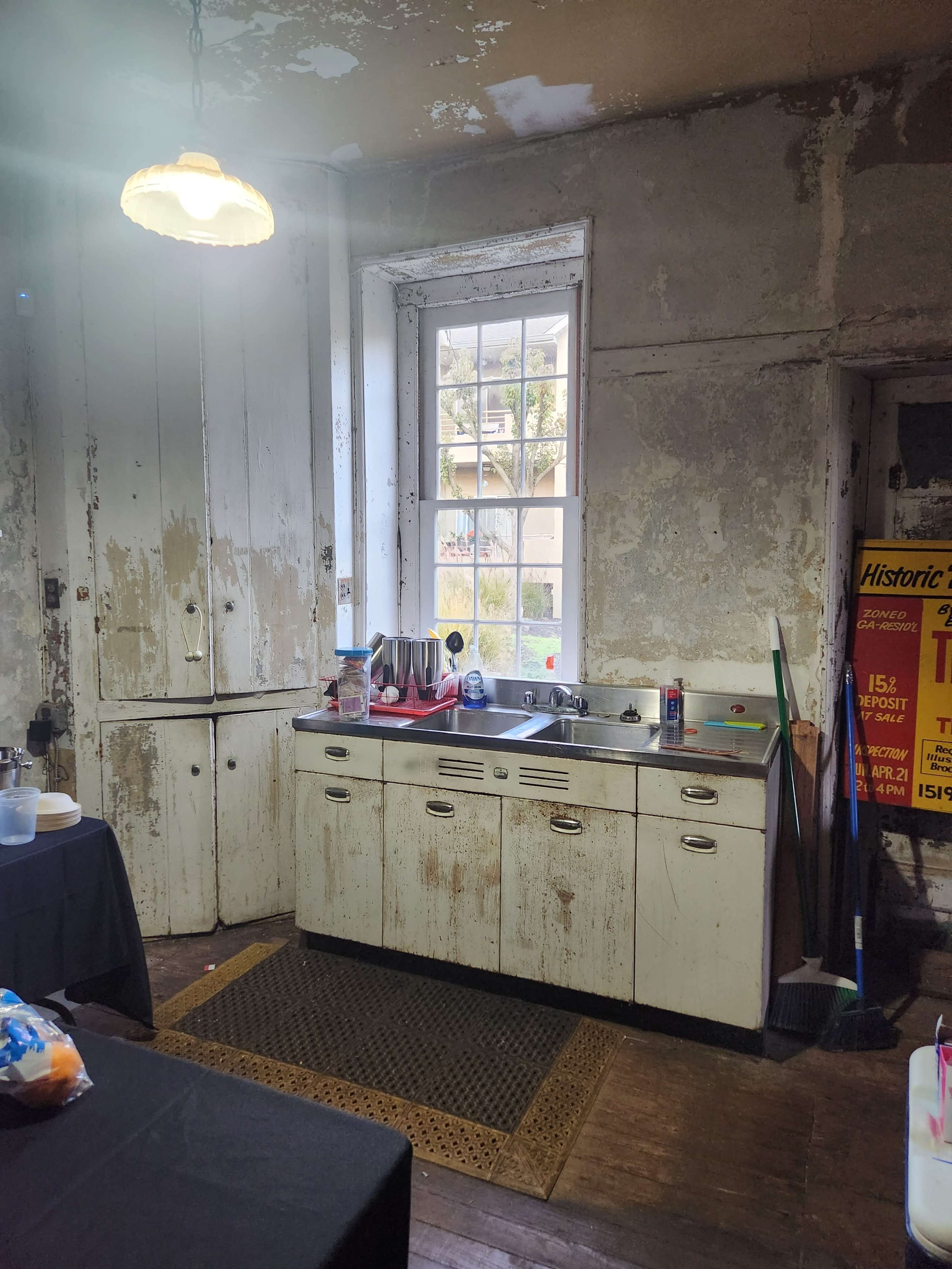 A weathered kitchen with a vintage sink, a window allowing natural light, and various cleaning tools nearby on the floor.