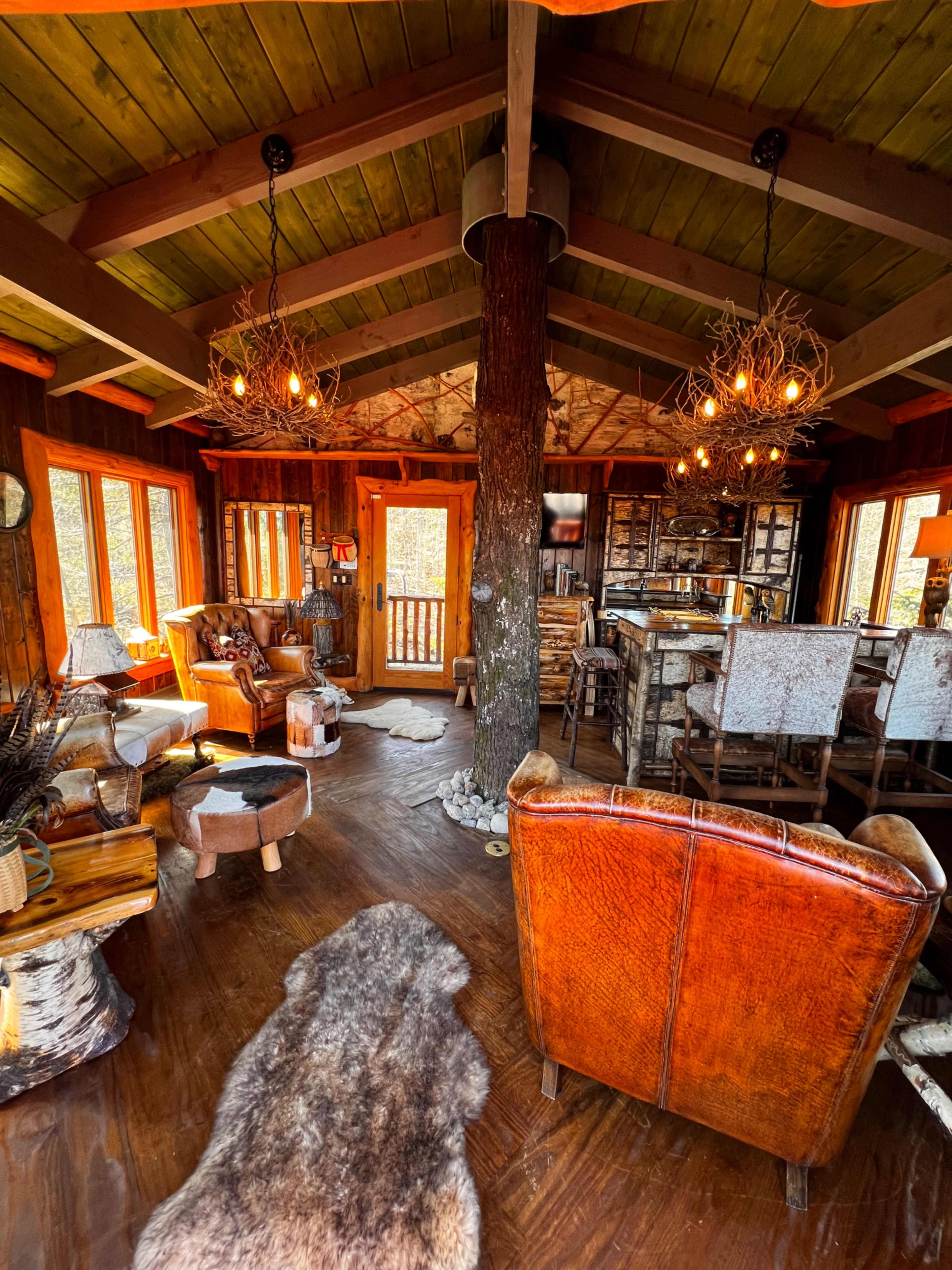 The interior of a rustic cabin features wooden walls, a central support pillar, cozy seating areas, and large windows allowing natural light.