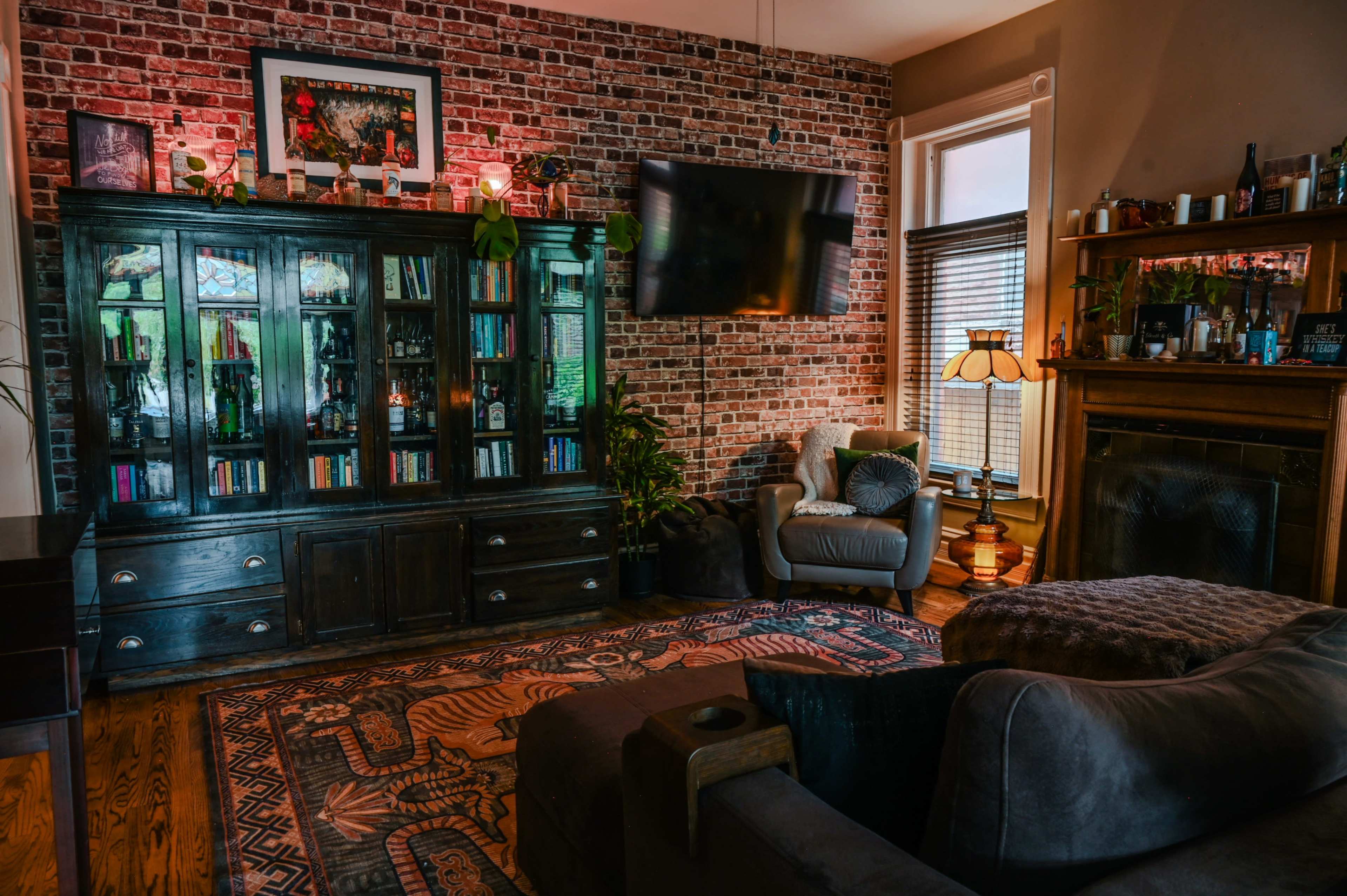 The scene features a cozy living room with a brick wall, a dark wooden cabinet filled with books and decor, a plush seating area, and a mounted television above the fireplace.