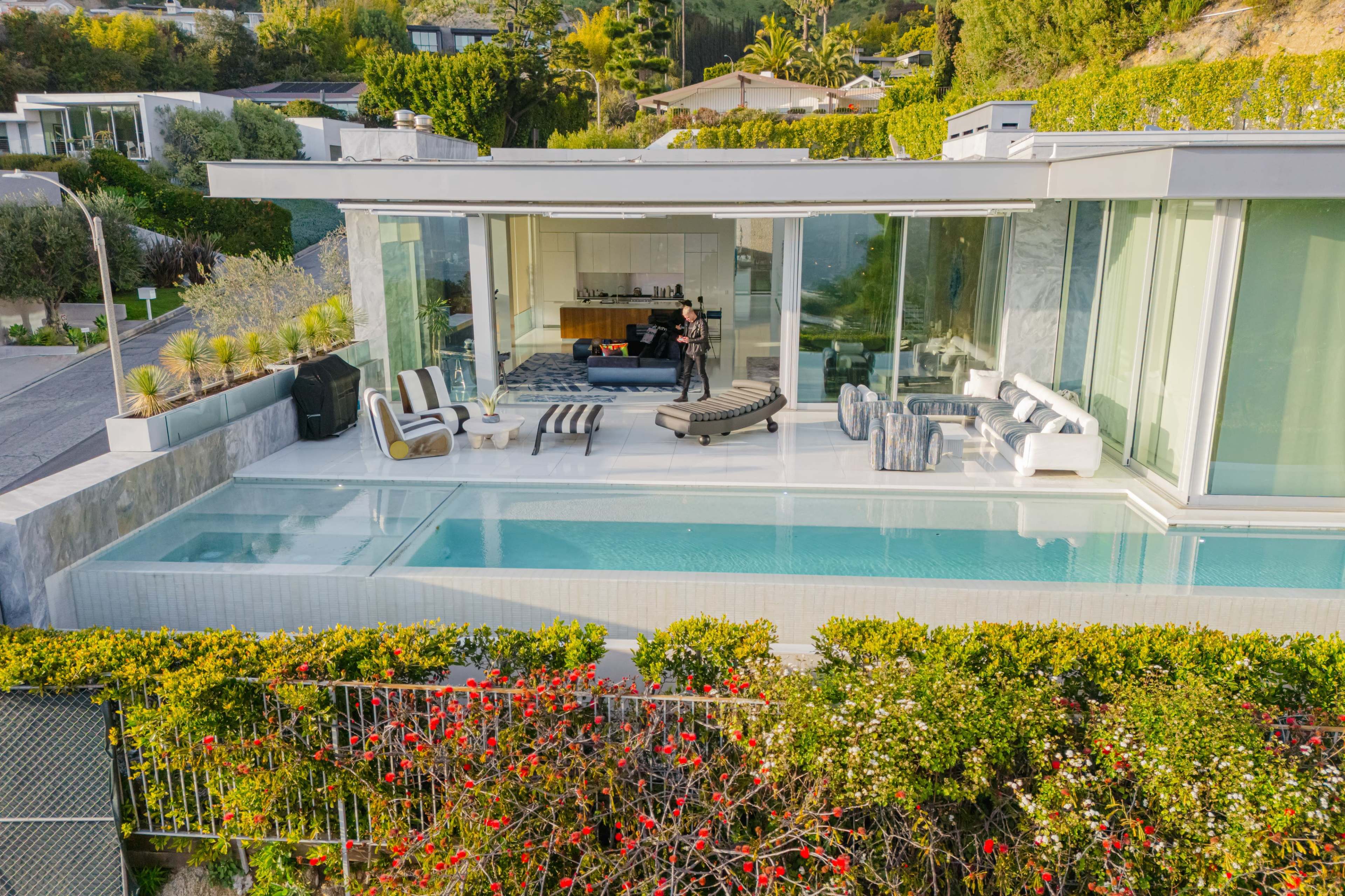 A modern house with large glass walls overlooks a pool and landscaped garden, featuring outdoor seating and a kitchen visible inside.