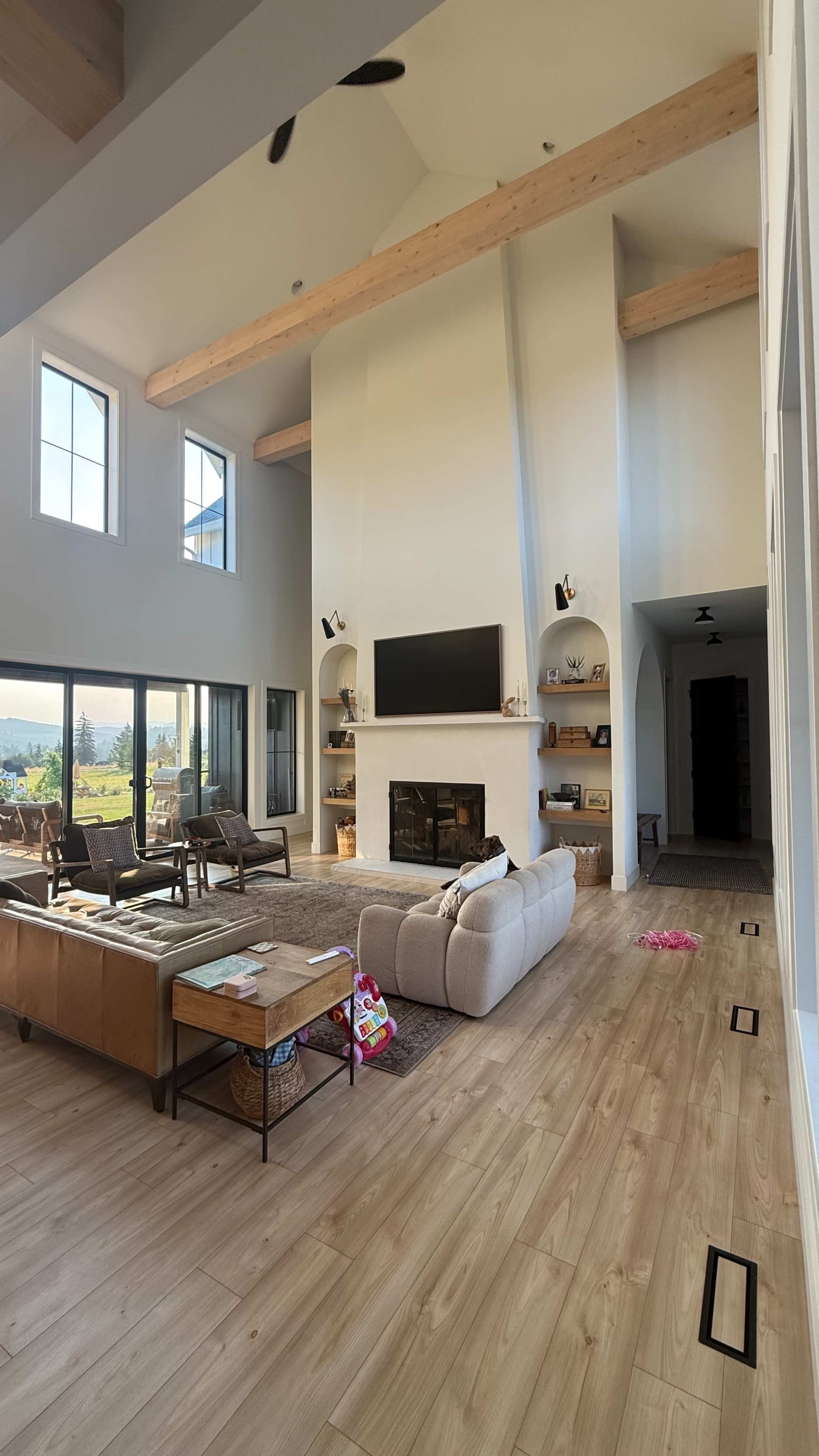 The image shows a spacious living room with a large sectional sofa, coffee table, and a flat-screen TV mounted on a white fireplace, surrounded by tall windows and wooden beams.
