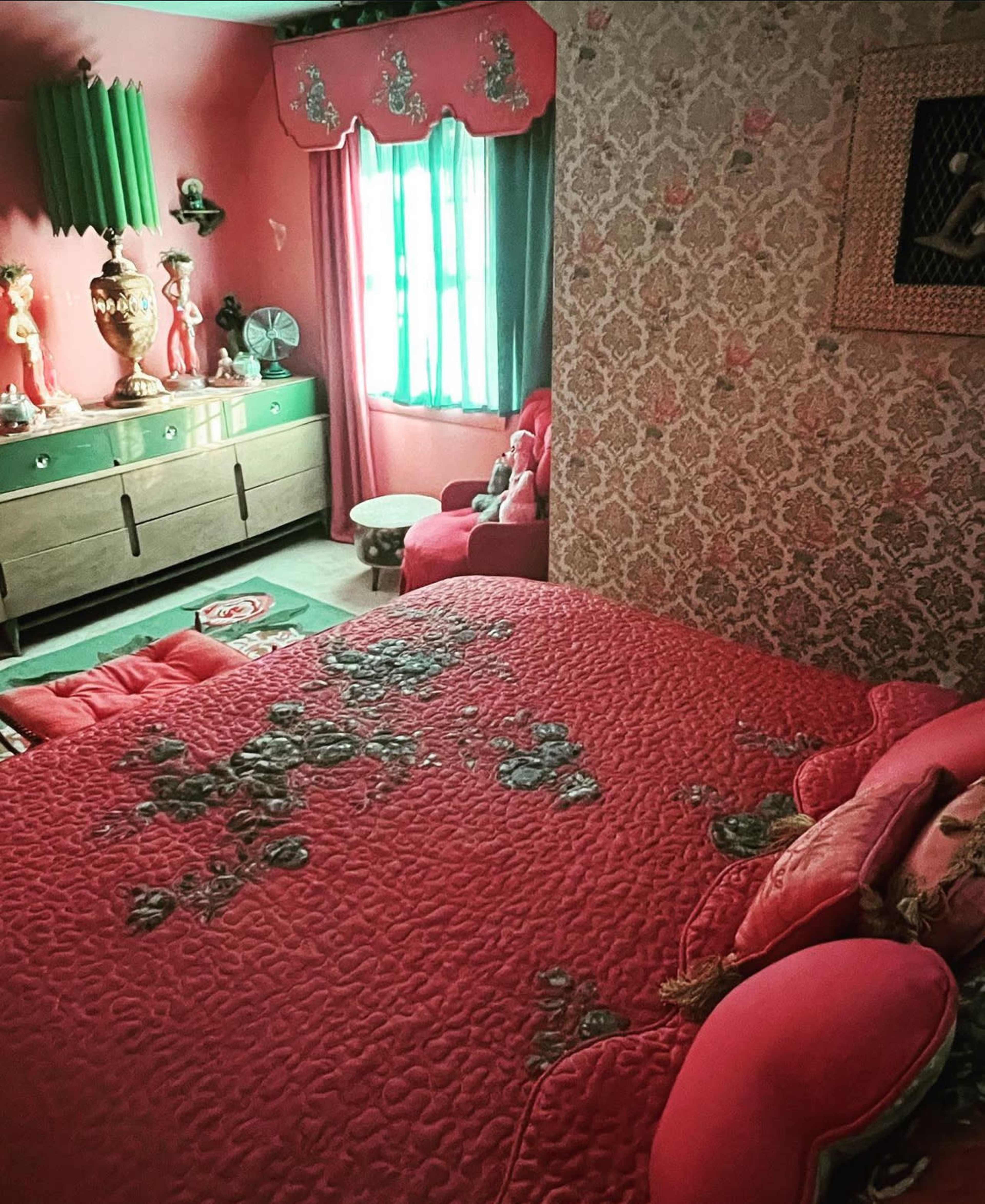 The image shows a vibrant bedroom with a red quilted bedspread, pink armchairs, and green curtains partially covering a window.