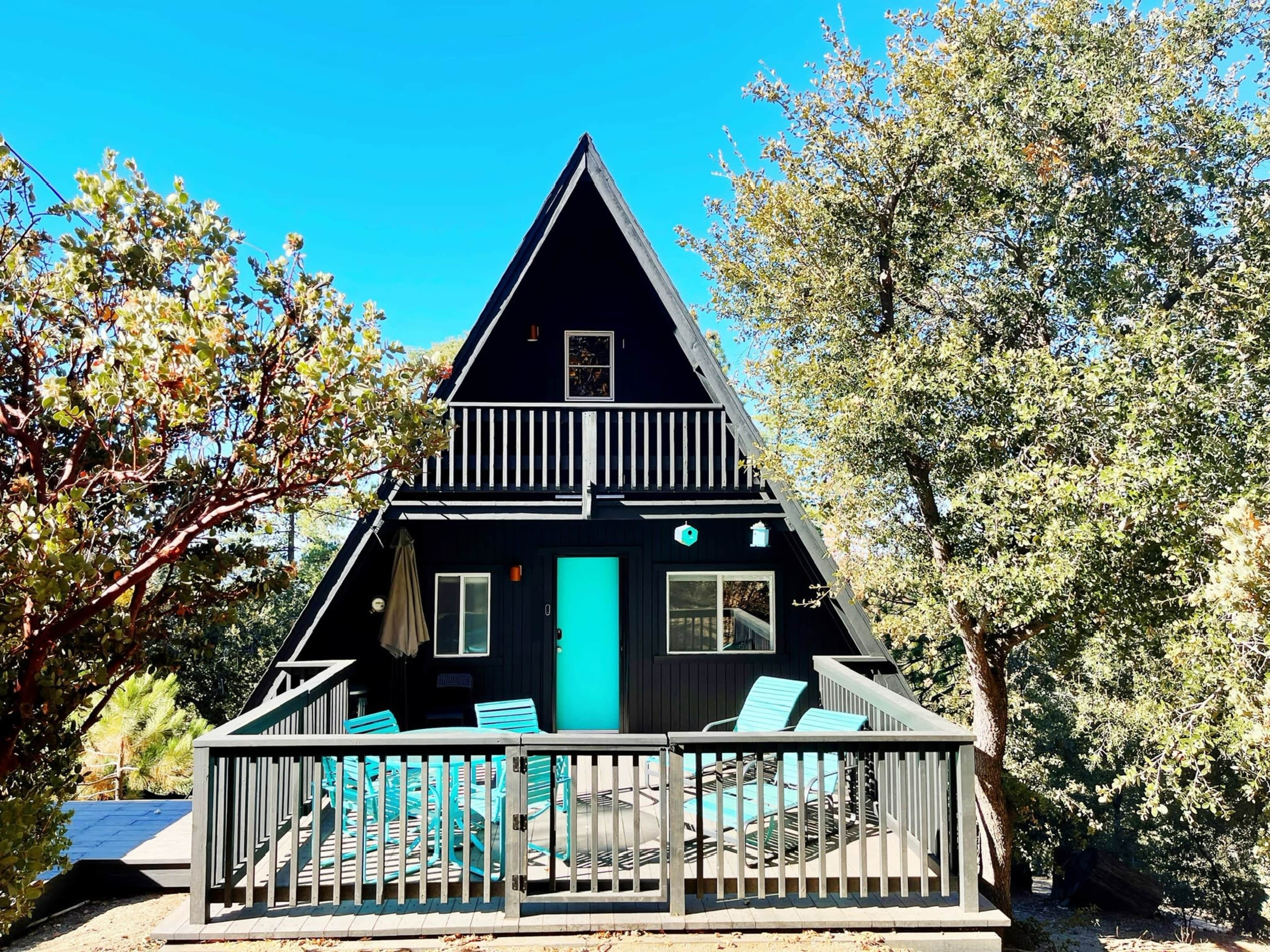A black A-frame house with a turquoise door and a surrounding deck is nestled among trees in a clear blue sky.