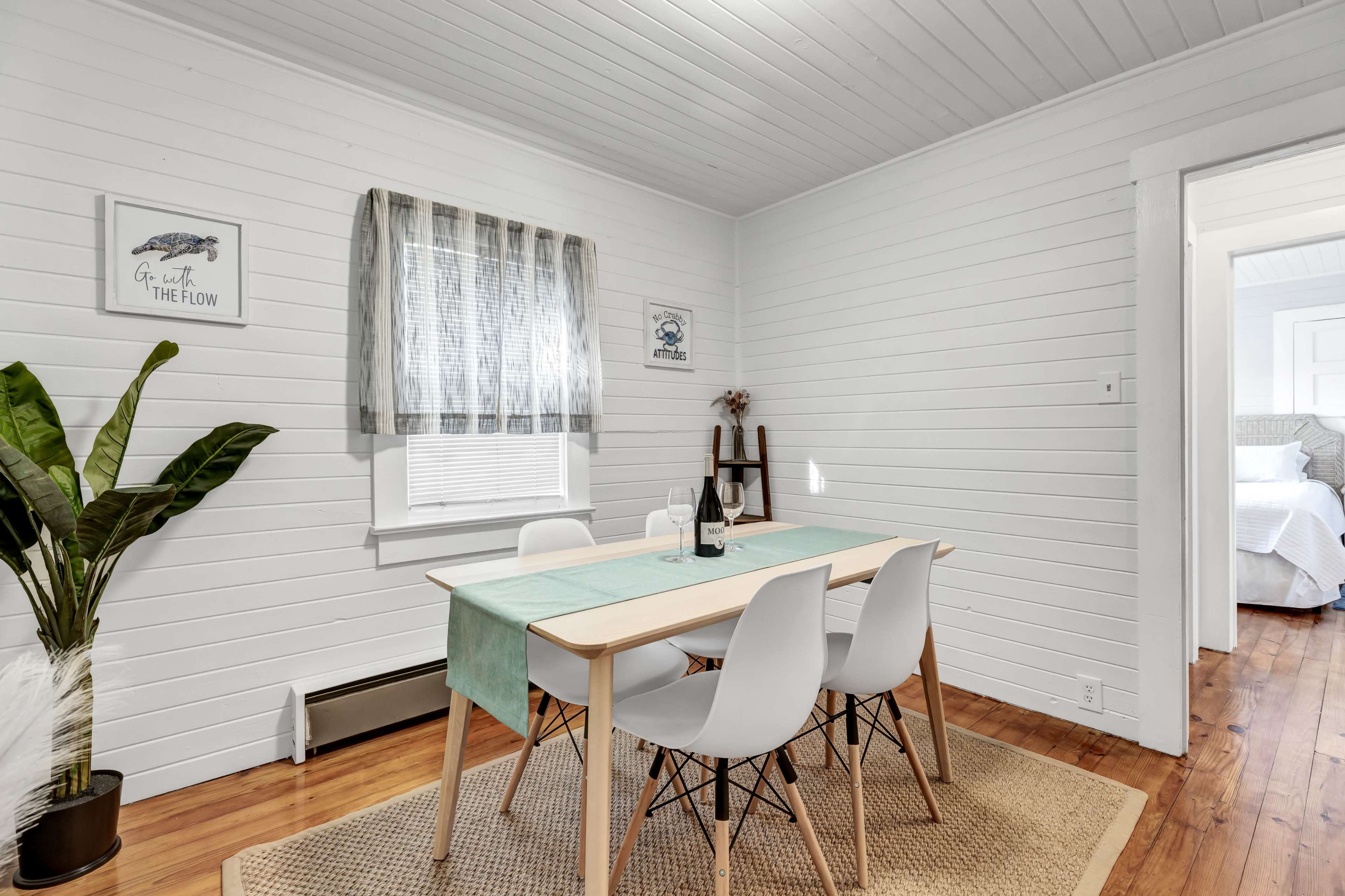 The image shows a bright dining area with a wooden table surrounded by white chairs, a plant in the corner, and light-colored walls.