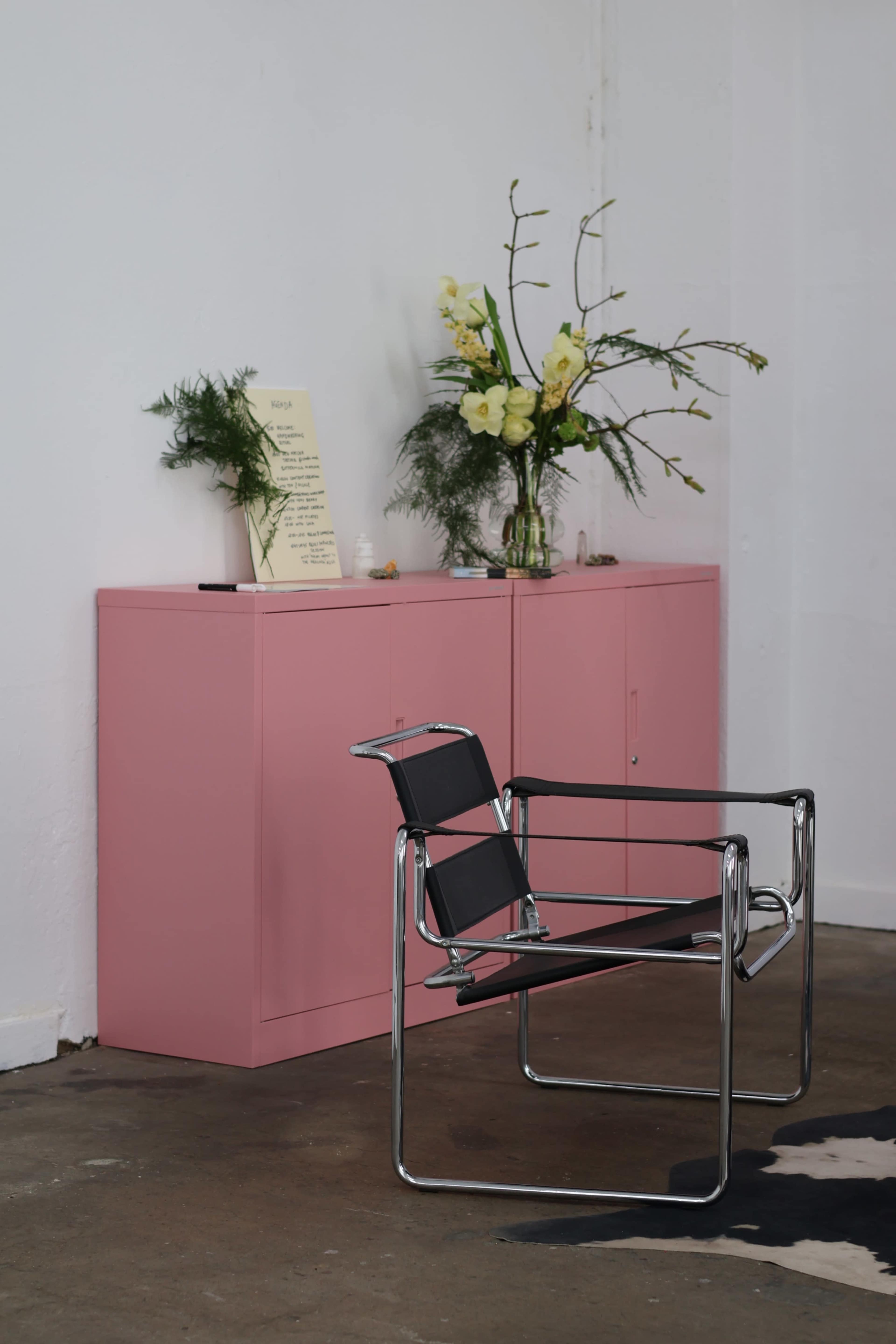 The image shows a pink cabinet against a white wall with a black chair in front of it, and a vase of yellow flowers on top of the cabinet.