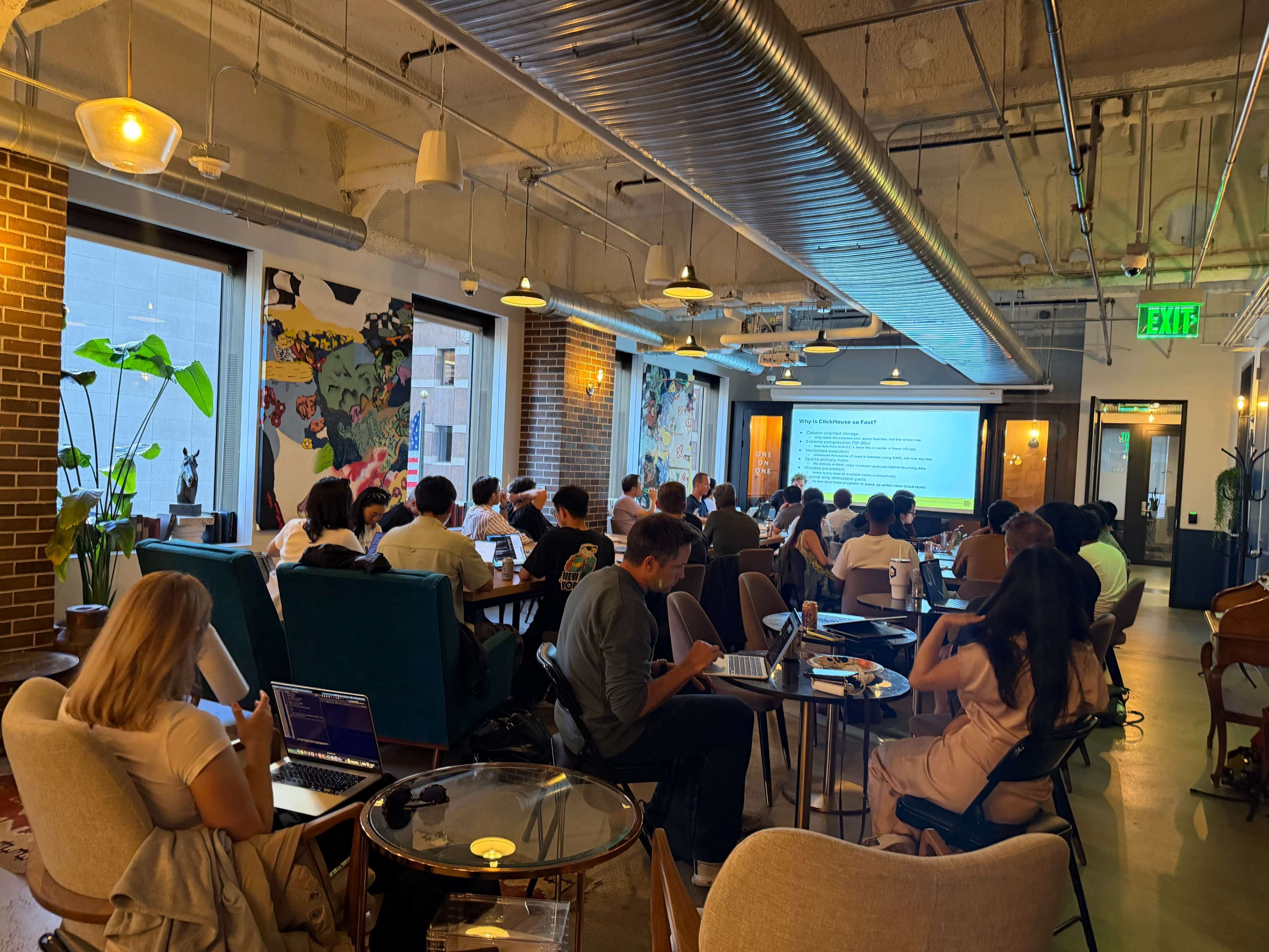 A group of people is seated at tables in a well-lit meeting space, attentively looking towards a screen displaying a presentation.